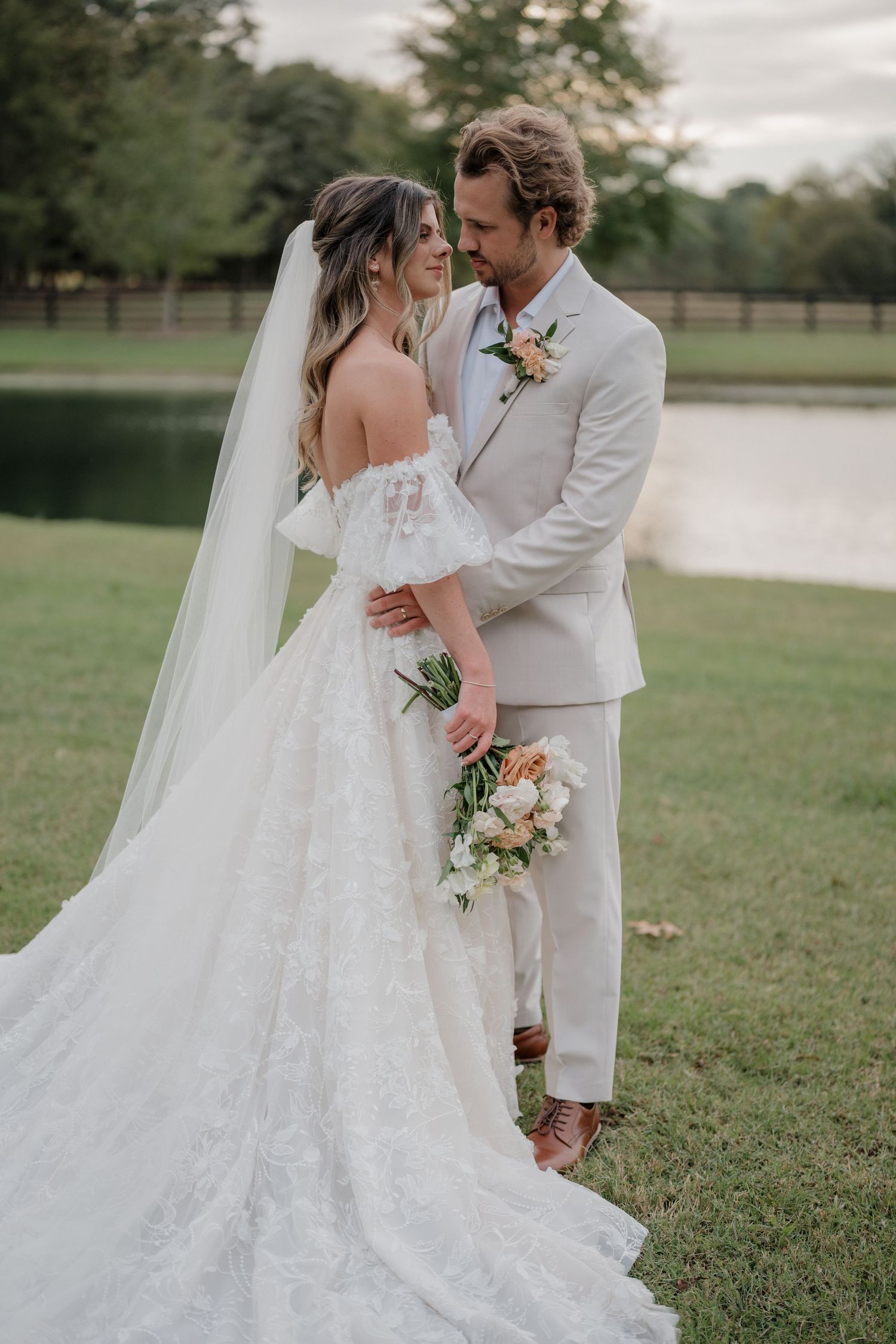 A romantic wedding couple embraces on a grassy lawn beside a lake, the bride wearing a flowing lace gown with a long veil.
