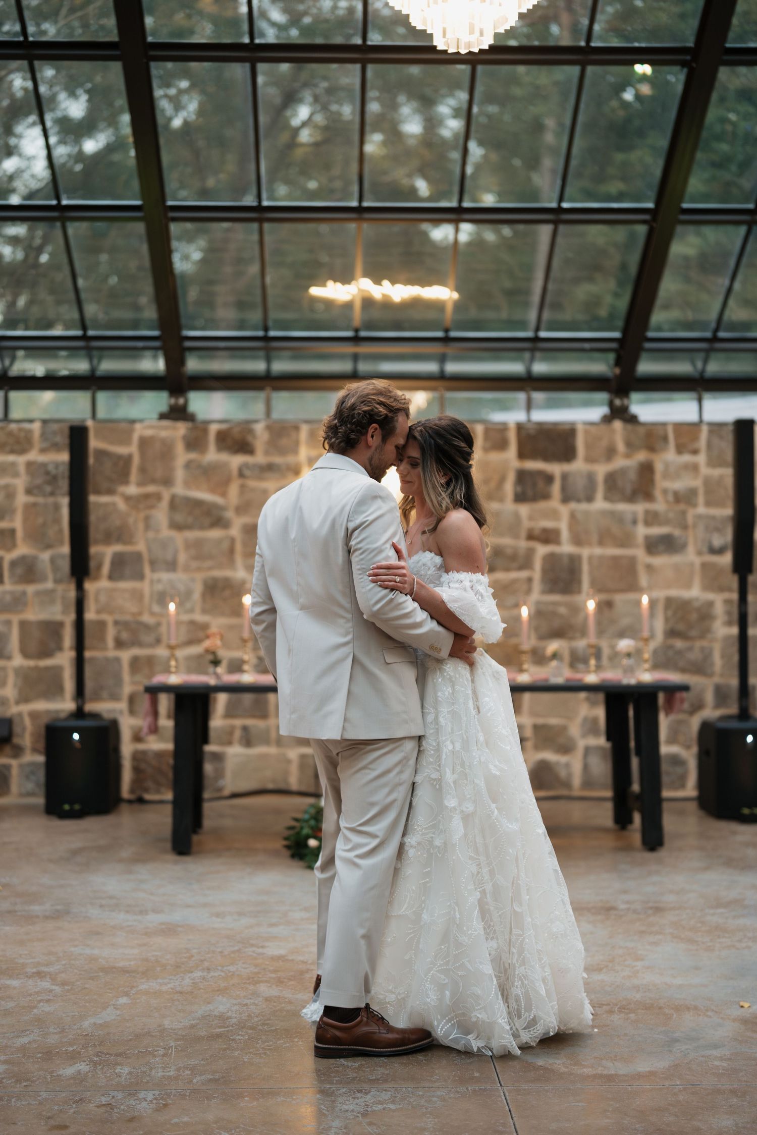 A romantic wedding couple dancing in an elegant venue with stone walls and chandeliers overhead.