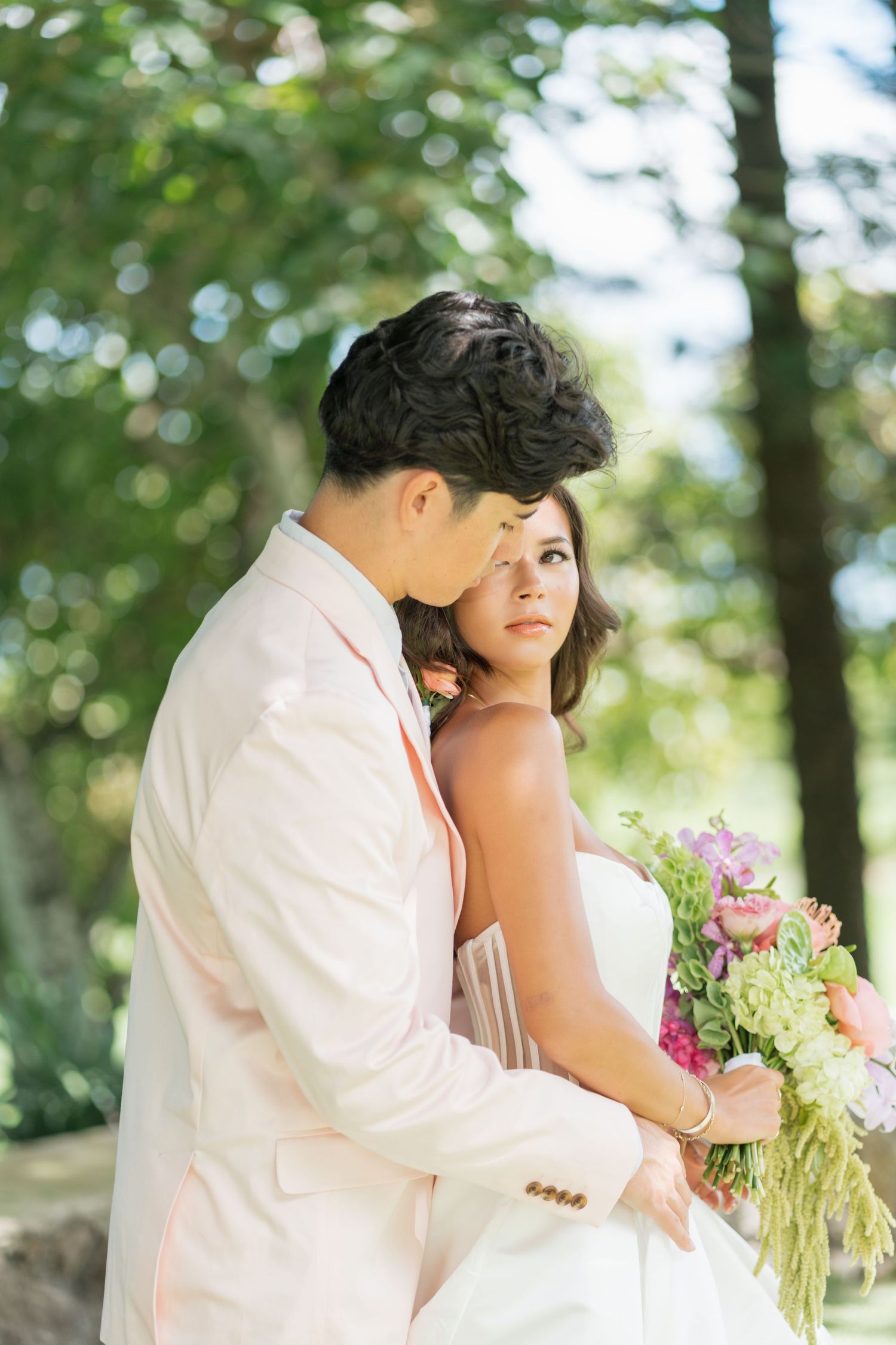 Asian bride and groom in white wedding attire embrace outdoors, bride holding colorful floral bouquet among lush green trees.