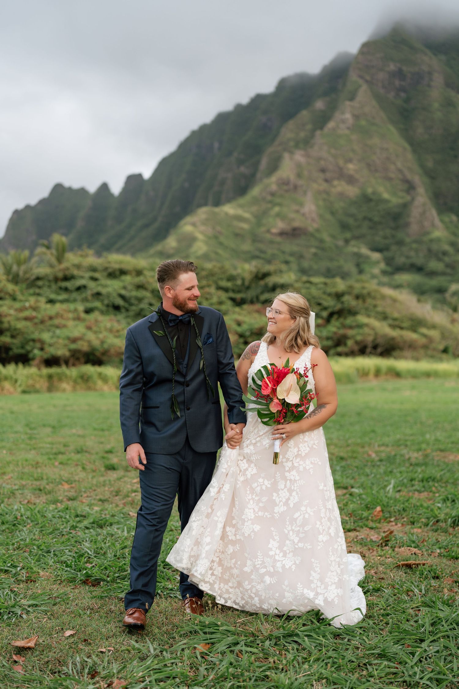 Bride and groom walk hand-in-hand in lush Hawaiian valley, she holds tropical bouquet, misty mountains backdrop.