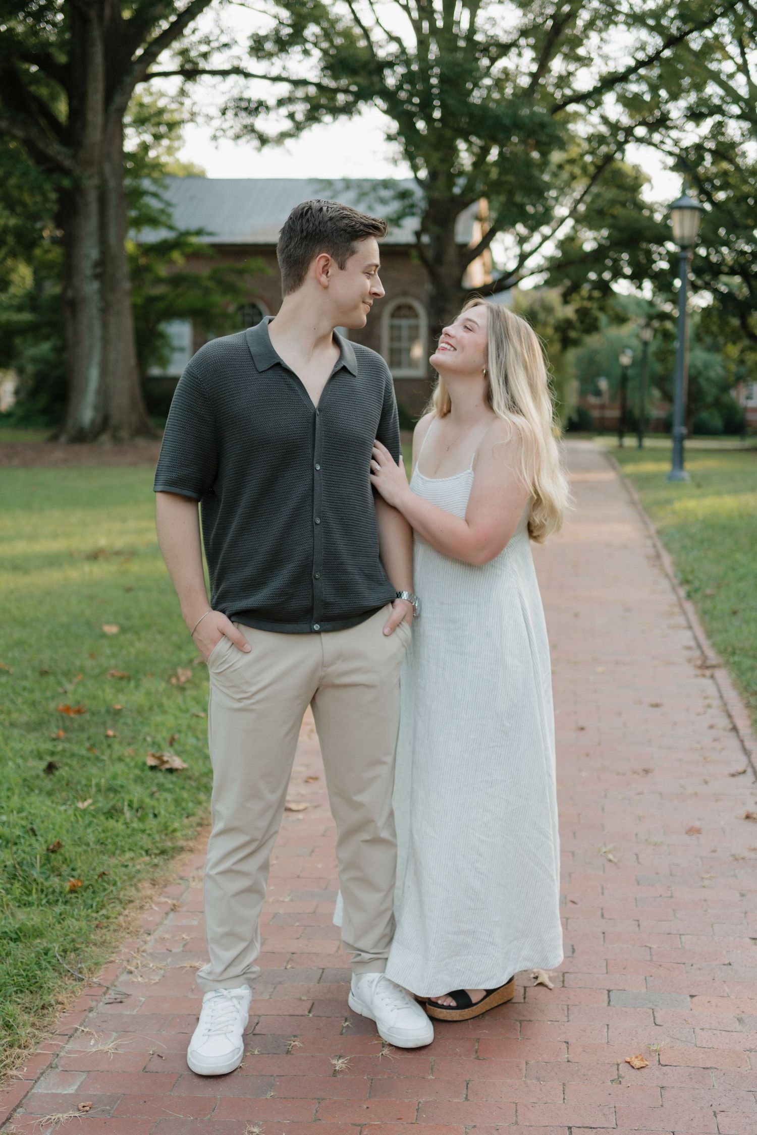 A couple in coordinated casual outfits share a romantic moment walking together on a brick pathway surrounded by trees.