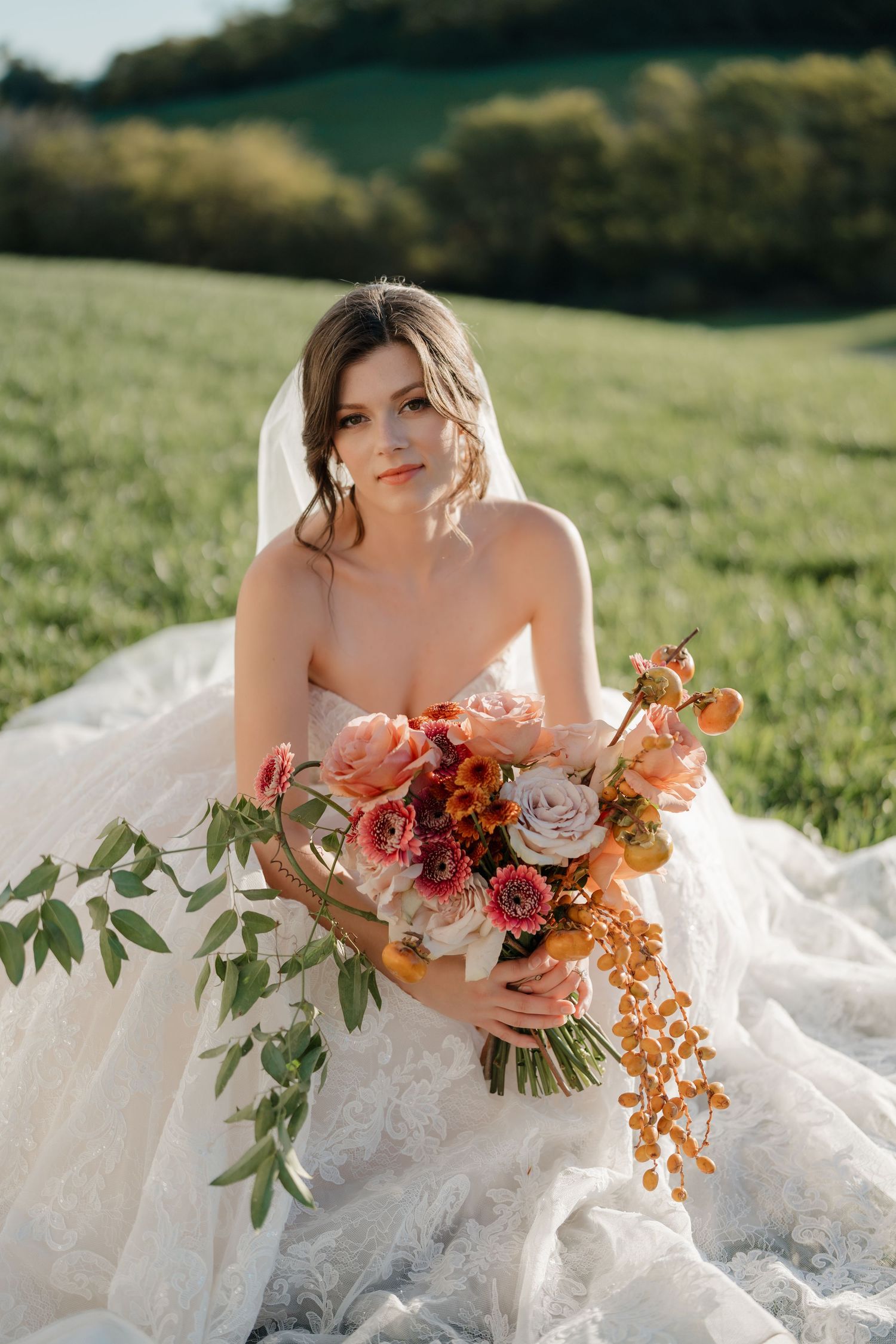 A wedding bouquet with coral and orange flowers rests on a flowing white lace dress in golden sunlight.