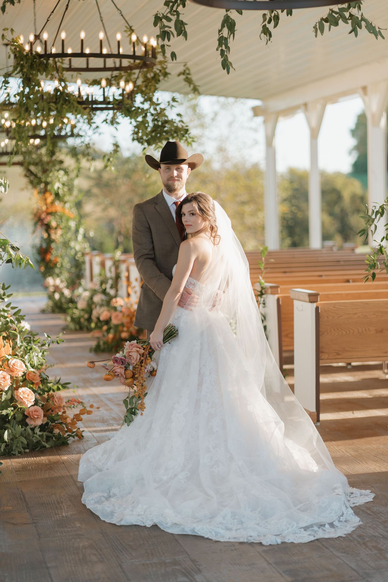 A romantic wedding scene in an outdoor chapel with wooden pews decorated with floral arrangements and greenery.