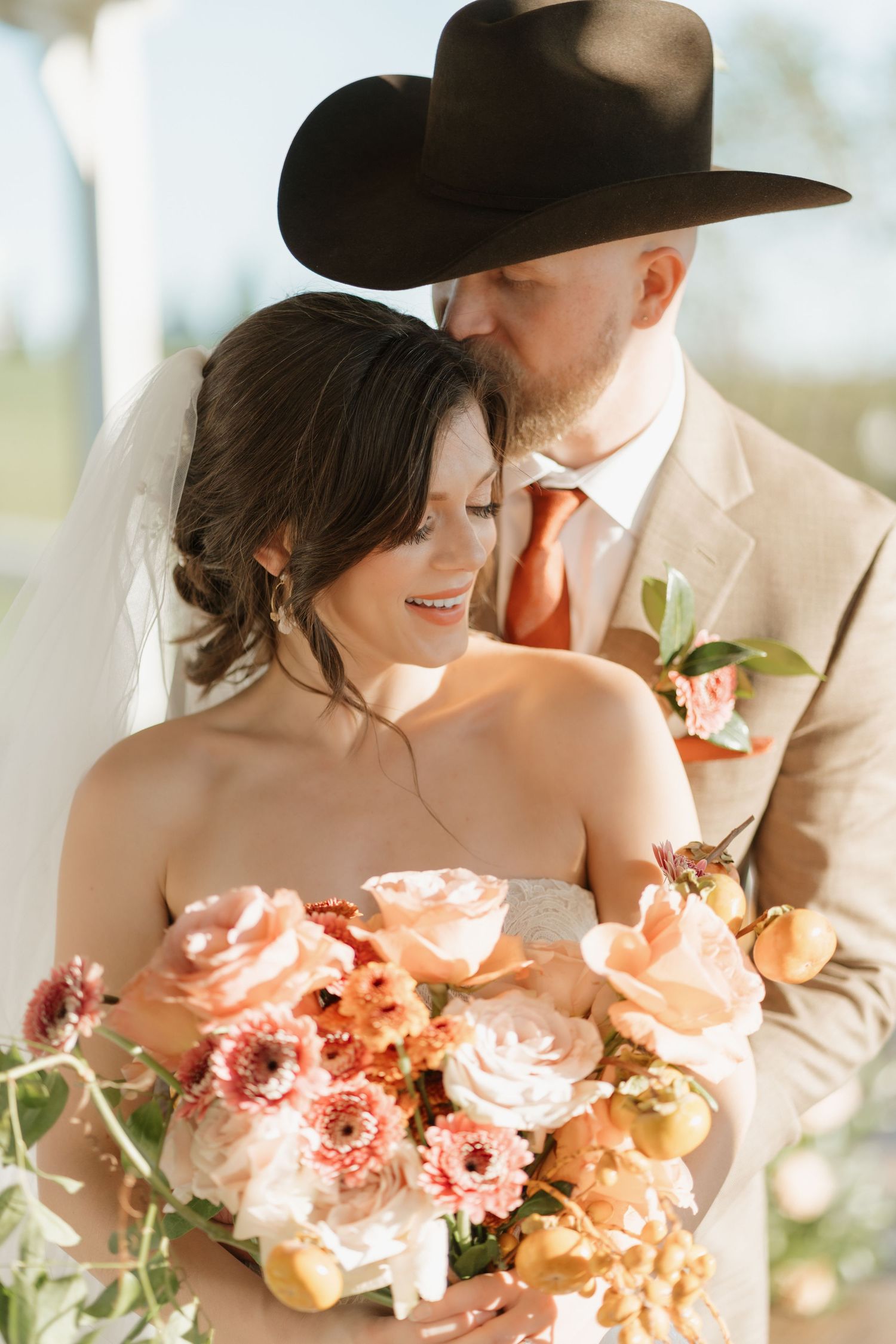 A romantic wedding couple in beige and black poses with a lush peach and coral floral bouquet.