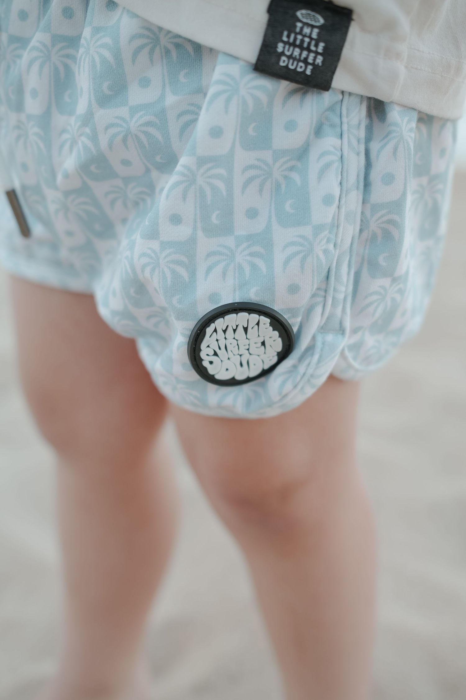 Close-up of toddler wearing light blue palm tree print shorts with a circular brand patch.