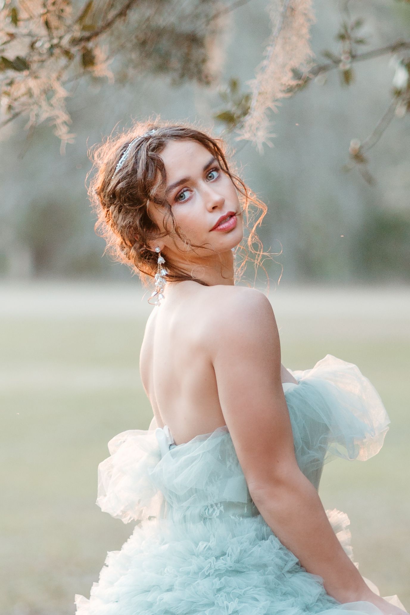 Young woman in a mint tulle gown poses outdoors under flowering branches in soft natural light.