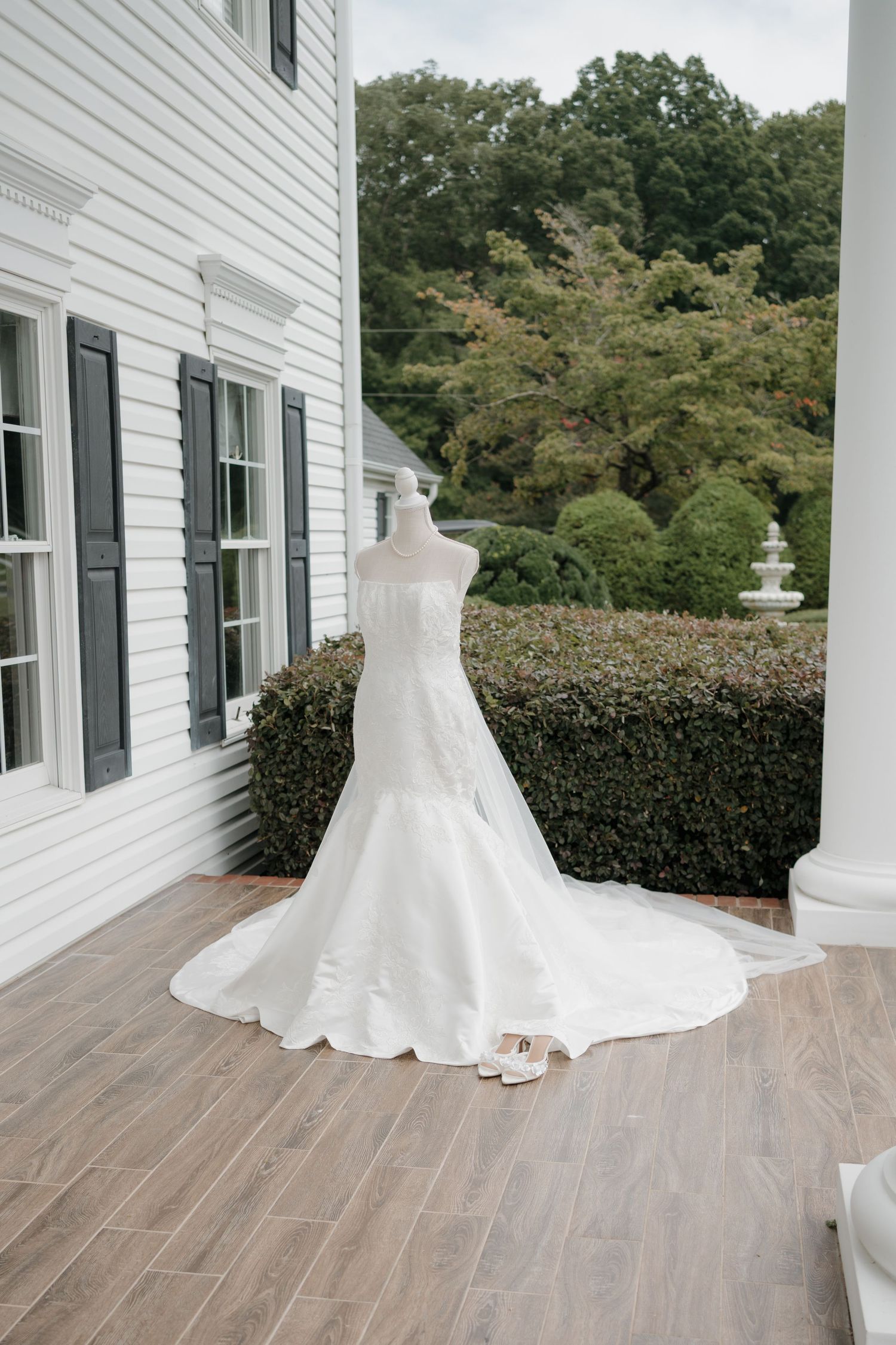 A classic white wedding dress is displayed on a form against white siding and black shutters on a porch.