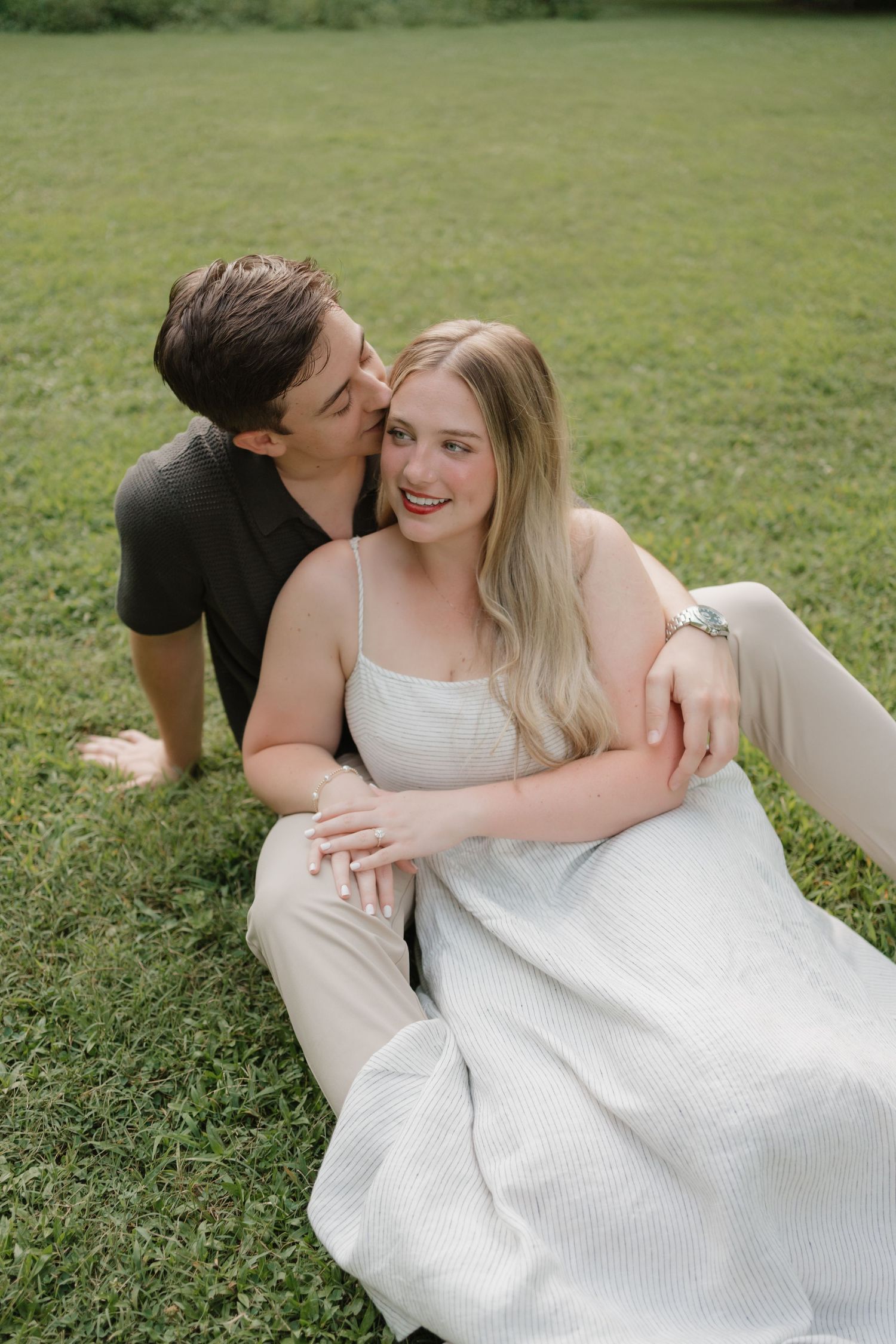A couple shares an intimate moment while relaxing on green grass, with her wearing a white dress and him in casual attire.