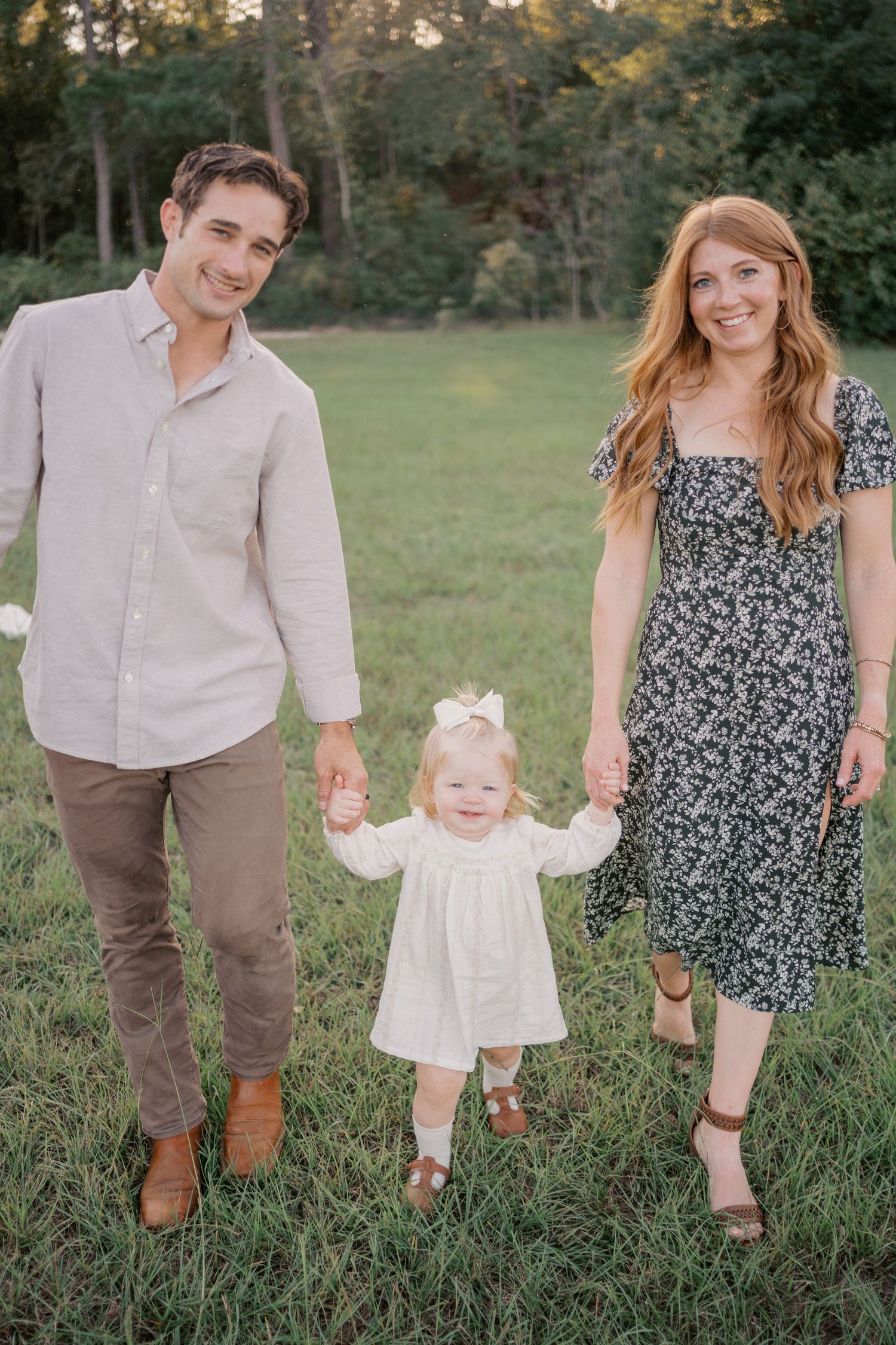 A couple and toddler in a white dress hold hands while walking through a grassy field during a family photoshoot at sunset.