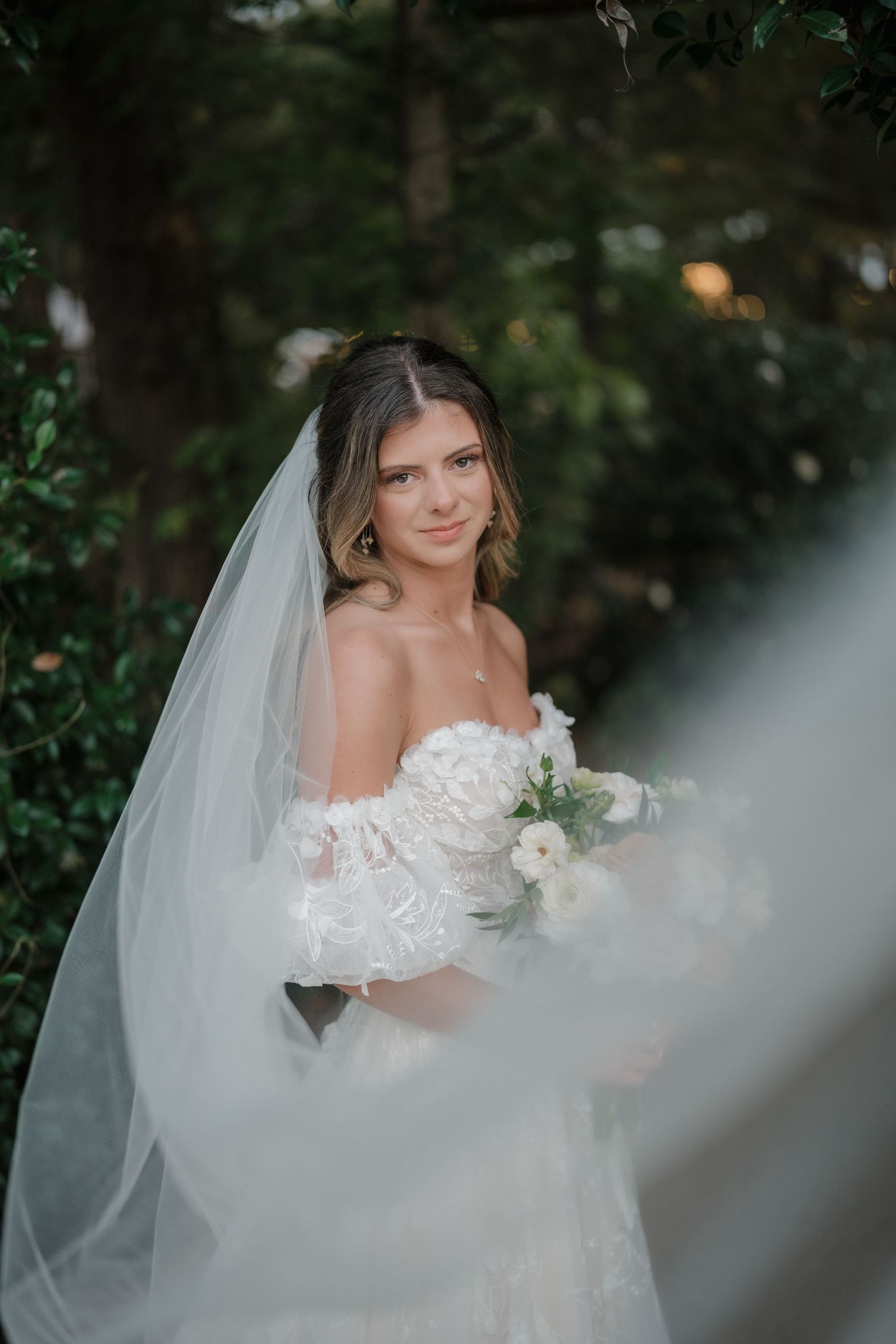 Ethereal bridal portrait through sheer veil with white floral bouquet in outdoor garden setting.