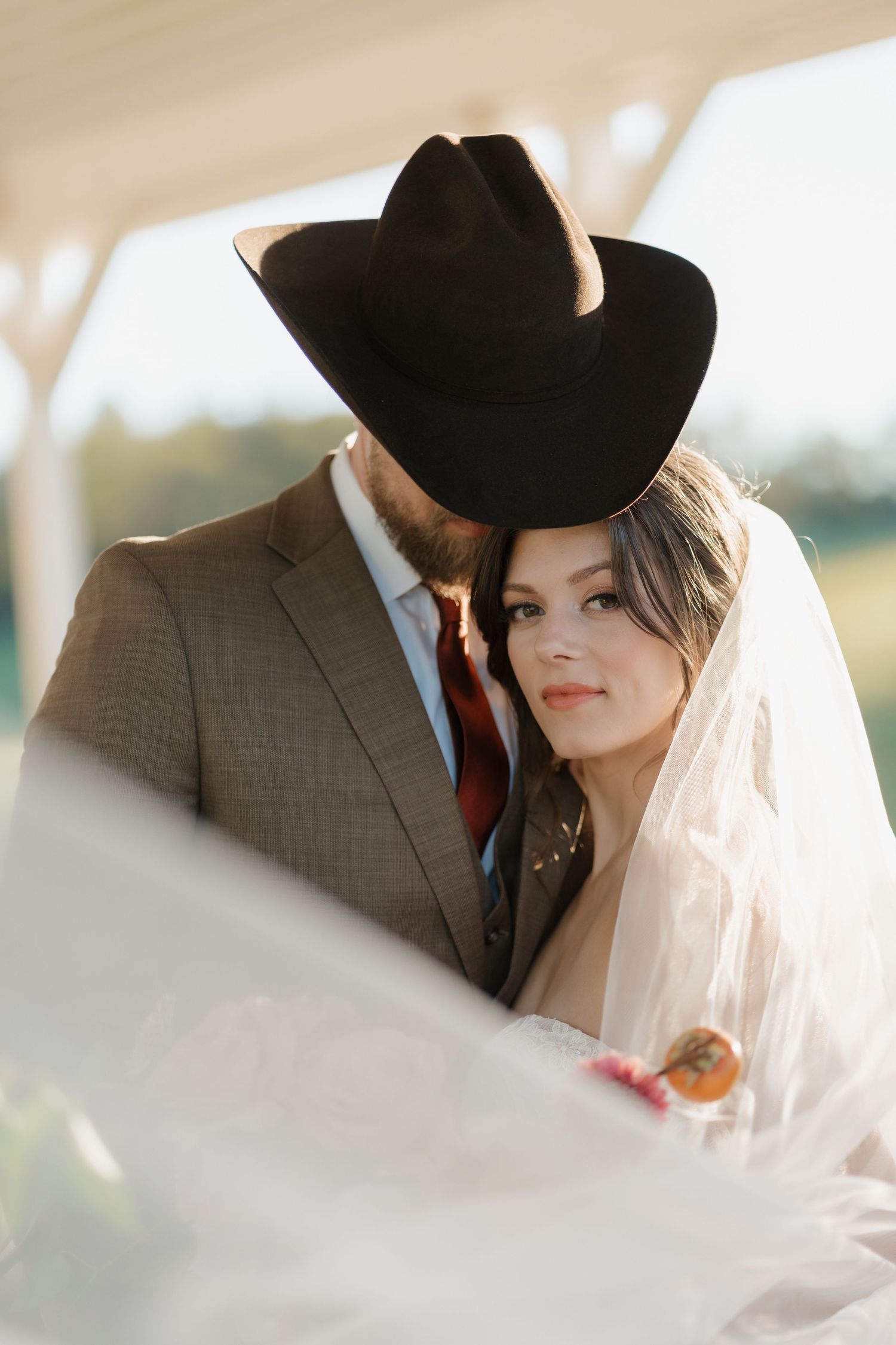 A romantic wedding couple in western attire share intimate moments under soft sunlight through a flowing white veil.