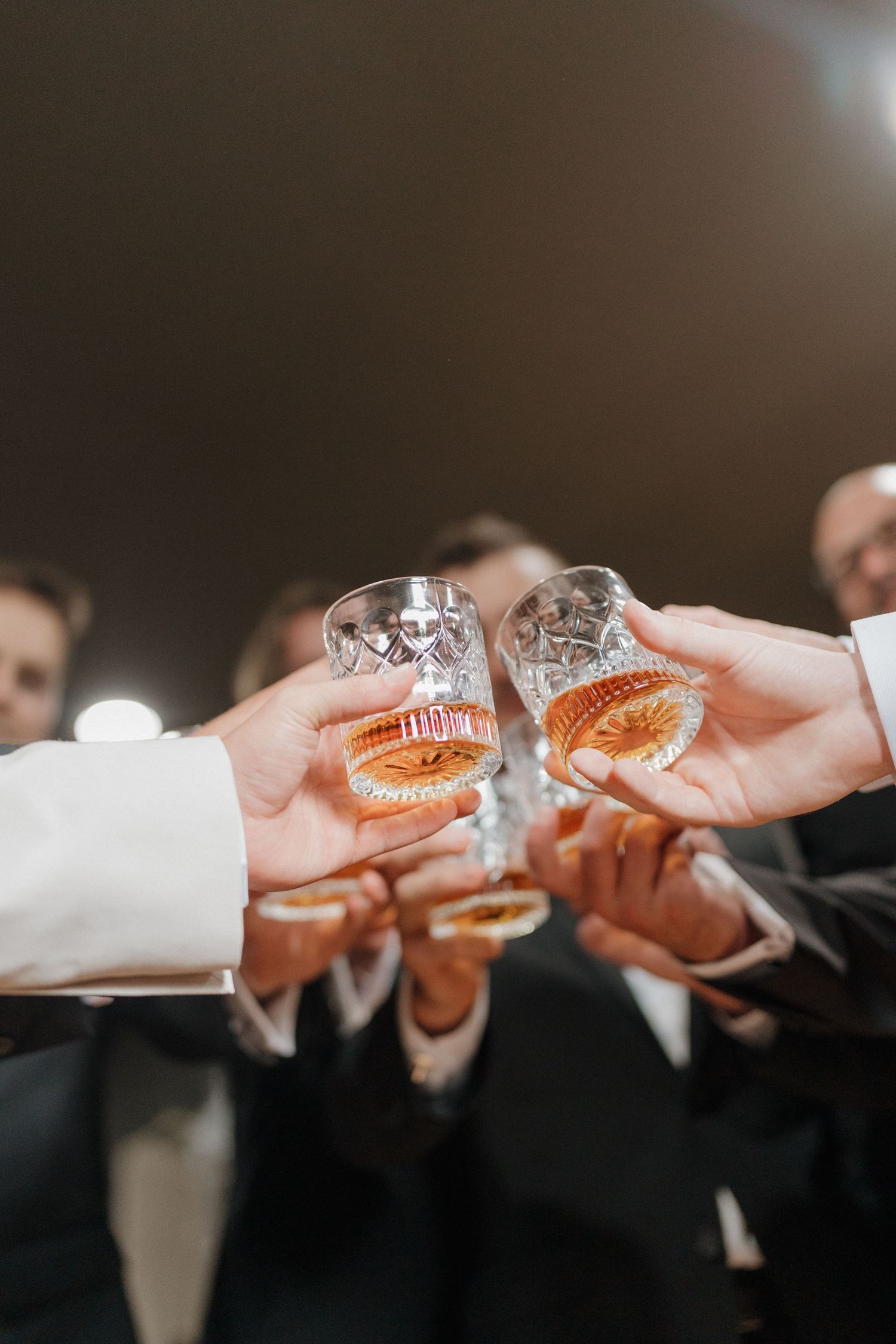 Several hands in formal attire raise crystal glasses filled with whiskey or bourbon for a celebratory toast.