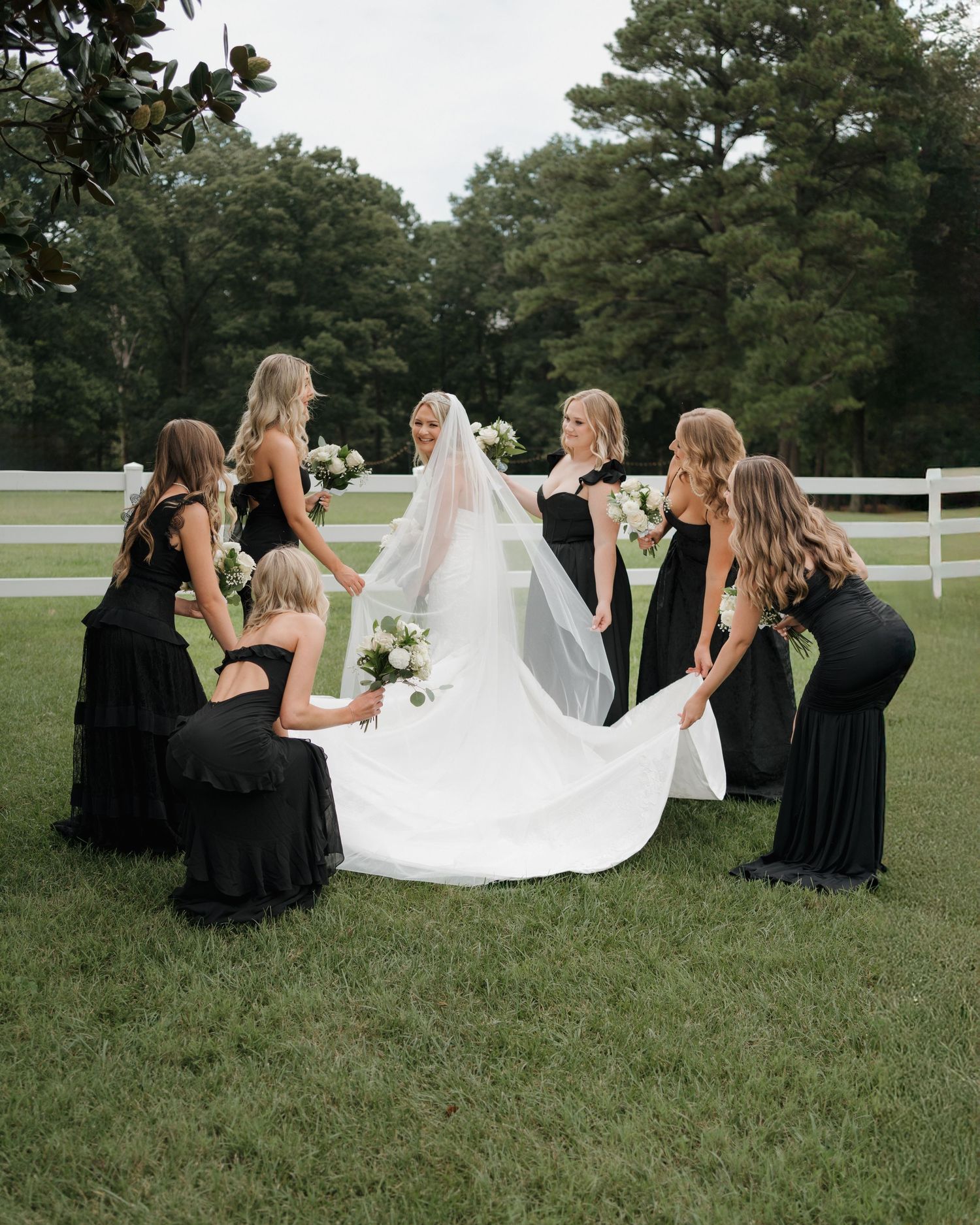 A group in elegant black dresses help arrange a flowing white wedding gown train on a grassy lawn with pine trees.