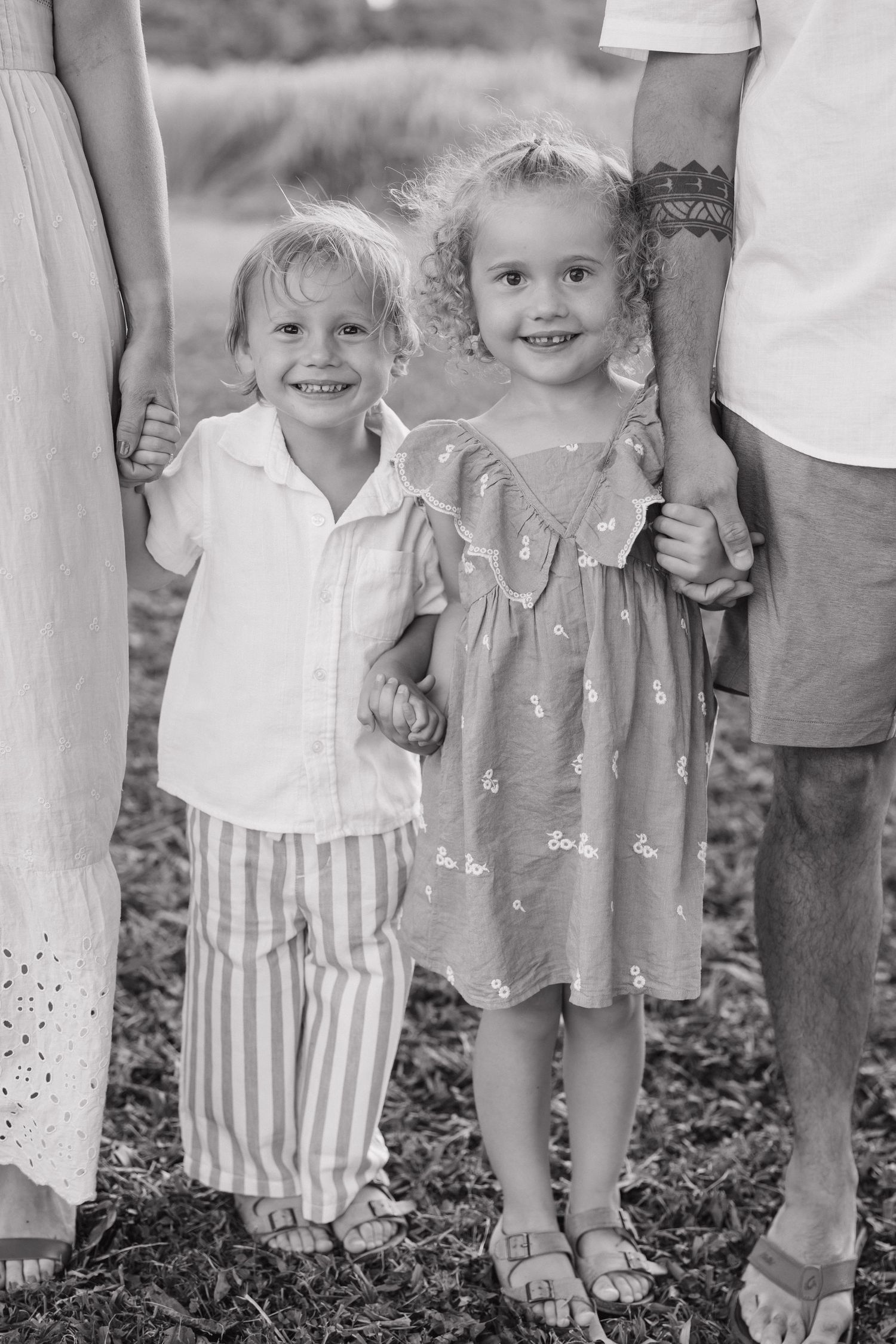 Black and white photo of two smiling young children holding hands with parents outdoors on grass.