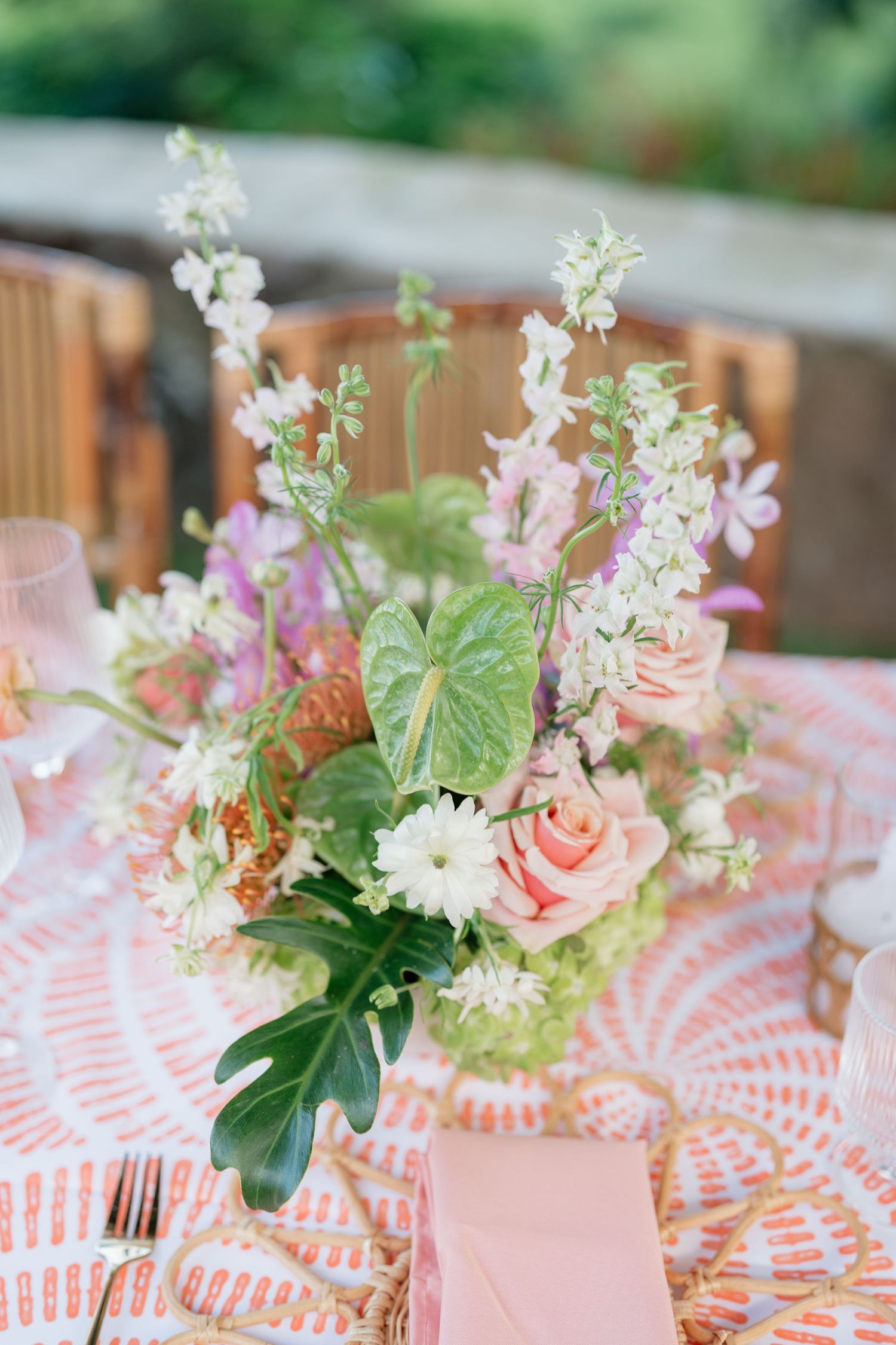 Elegant floral centerpiece with peach roses, green anthurium, and white blooms on a pink patterned tablecloth at an outdoor event.