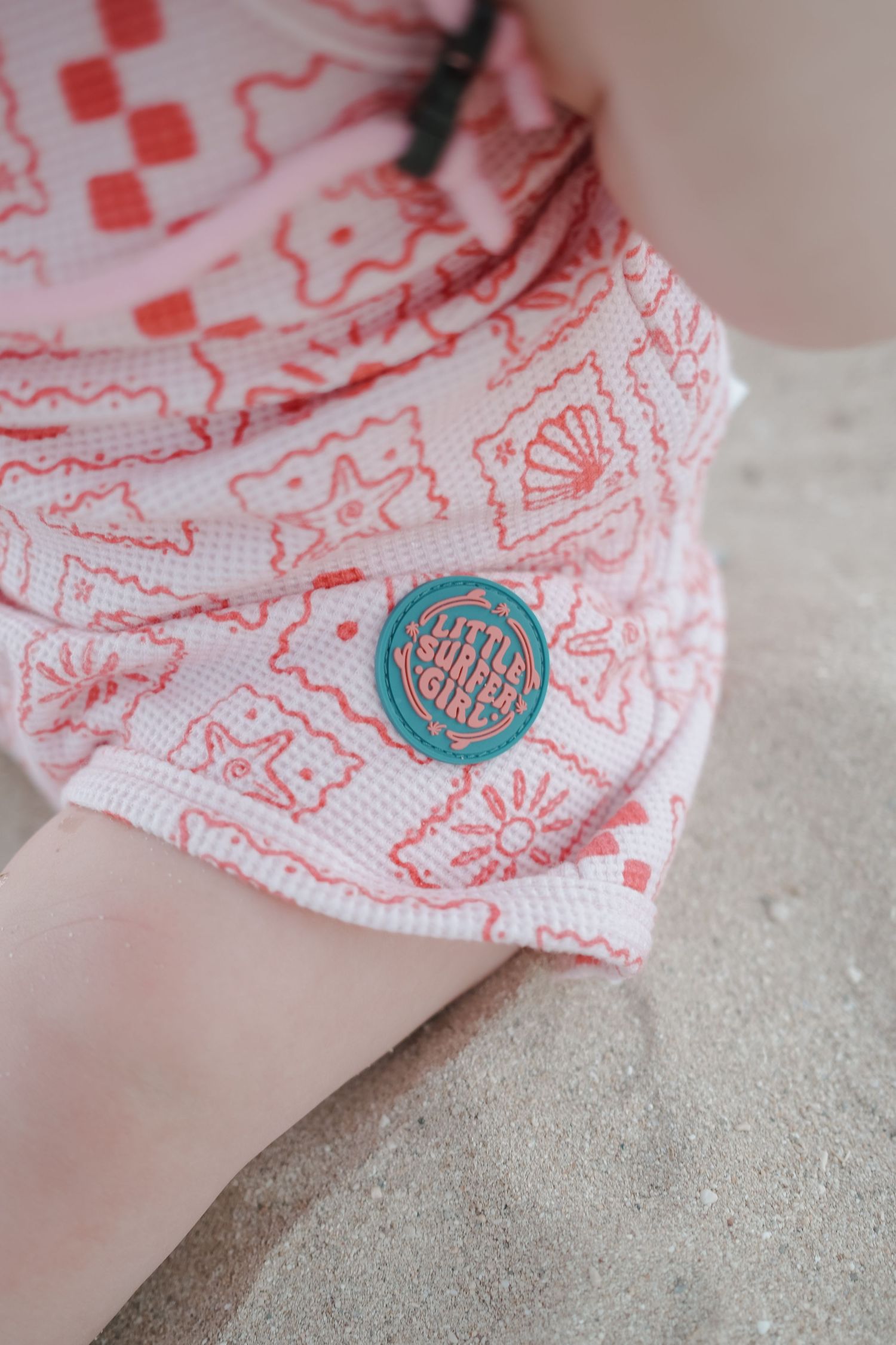 Pink patterned Little Surfer Girls shorts with teal logo patch, worn at the beach.
