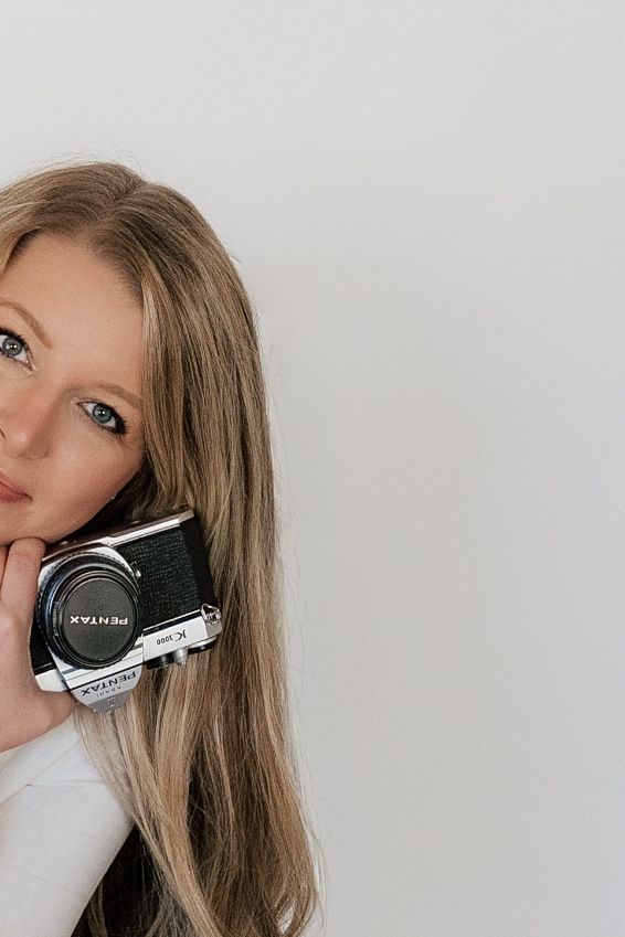A person with long blonde hair poses against a white background while holding a vintage film camera.