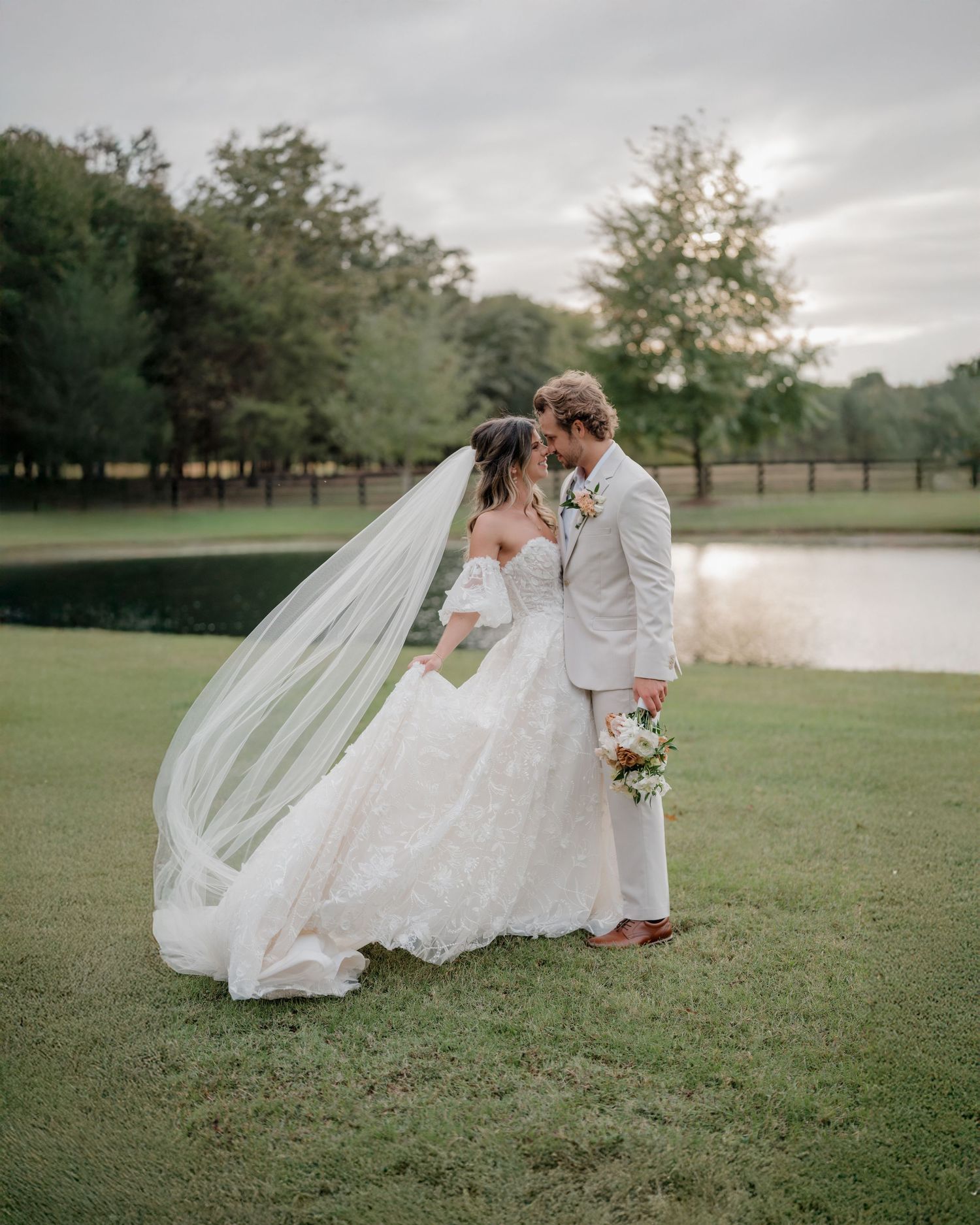 Wedding couple shares intimate moments on grassy lawn with flowing white dress and veil near lake.