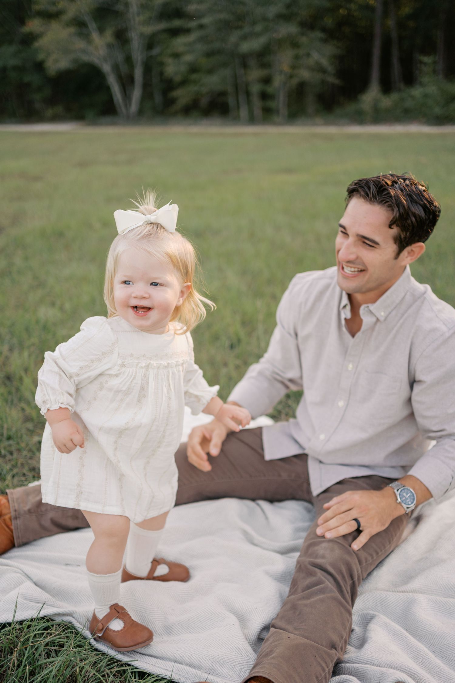 A small child in a white dress and bow plays on a picnic blanket outdoors.
