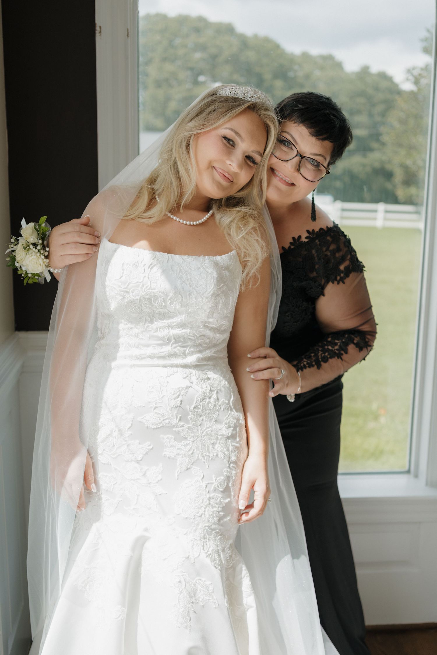A bride in a fitted lace wedding gown poses near a window while someone in black lace adjusts her dress.