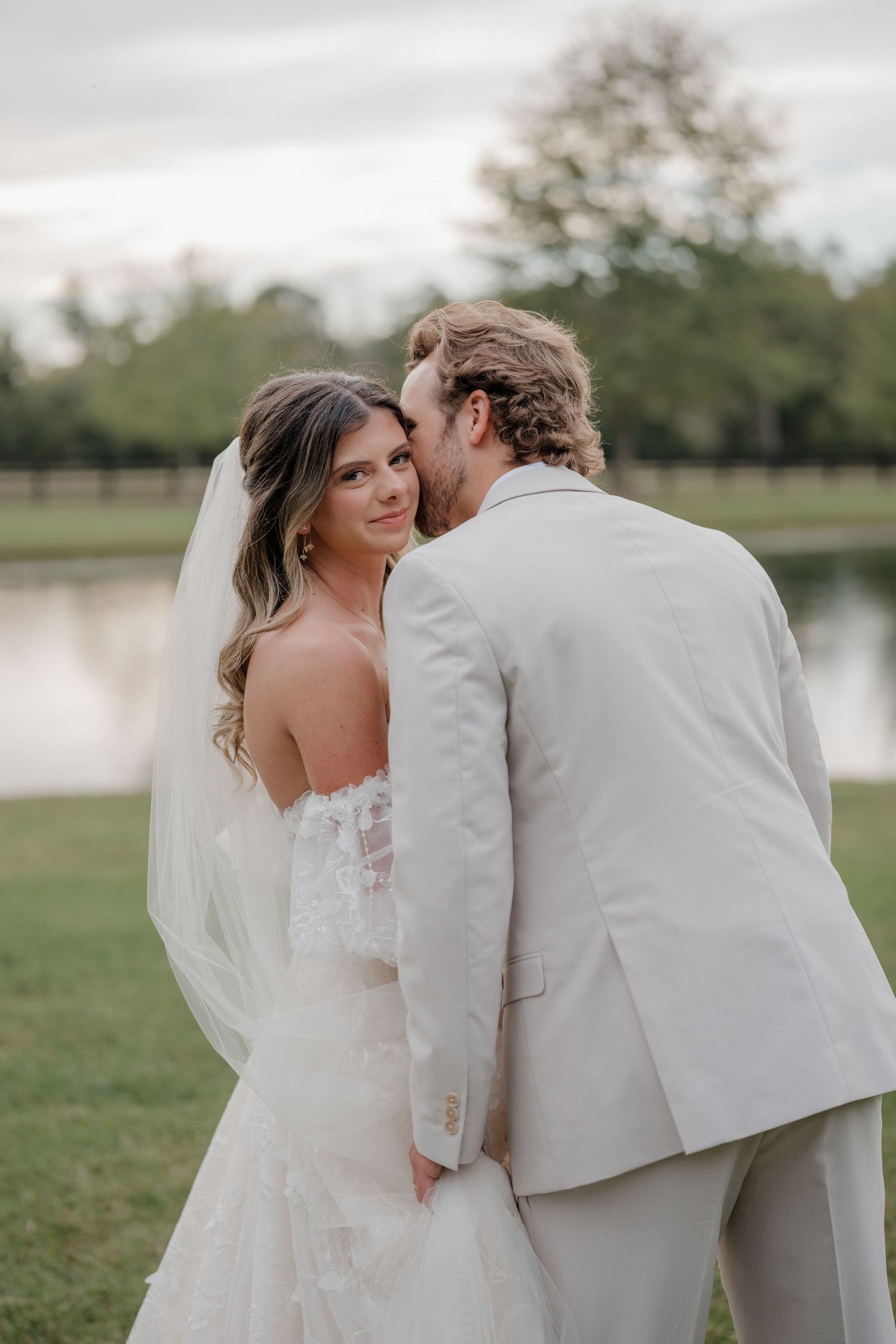 A romantic wedding couple shares an intimate moment outdoors by a lake in a natural setting.