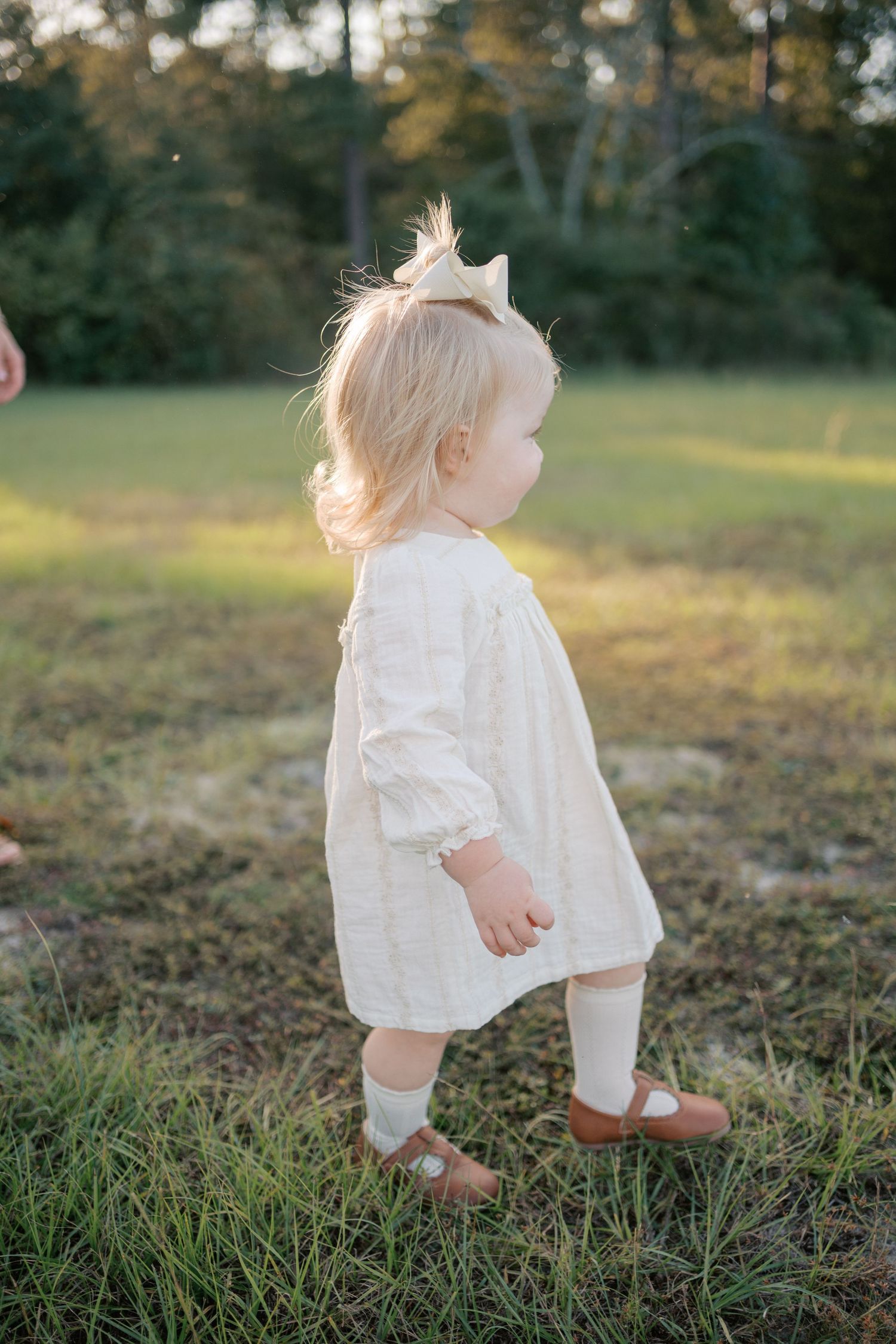 A small child in a white dress with ruffles walks through grassy field during golden hour.