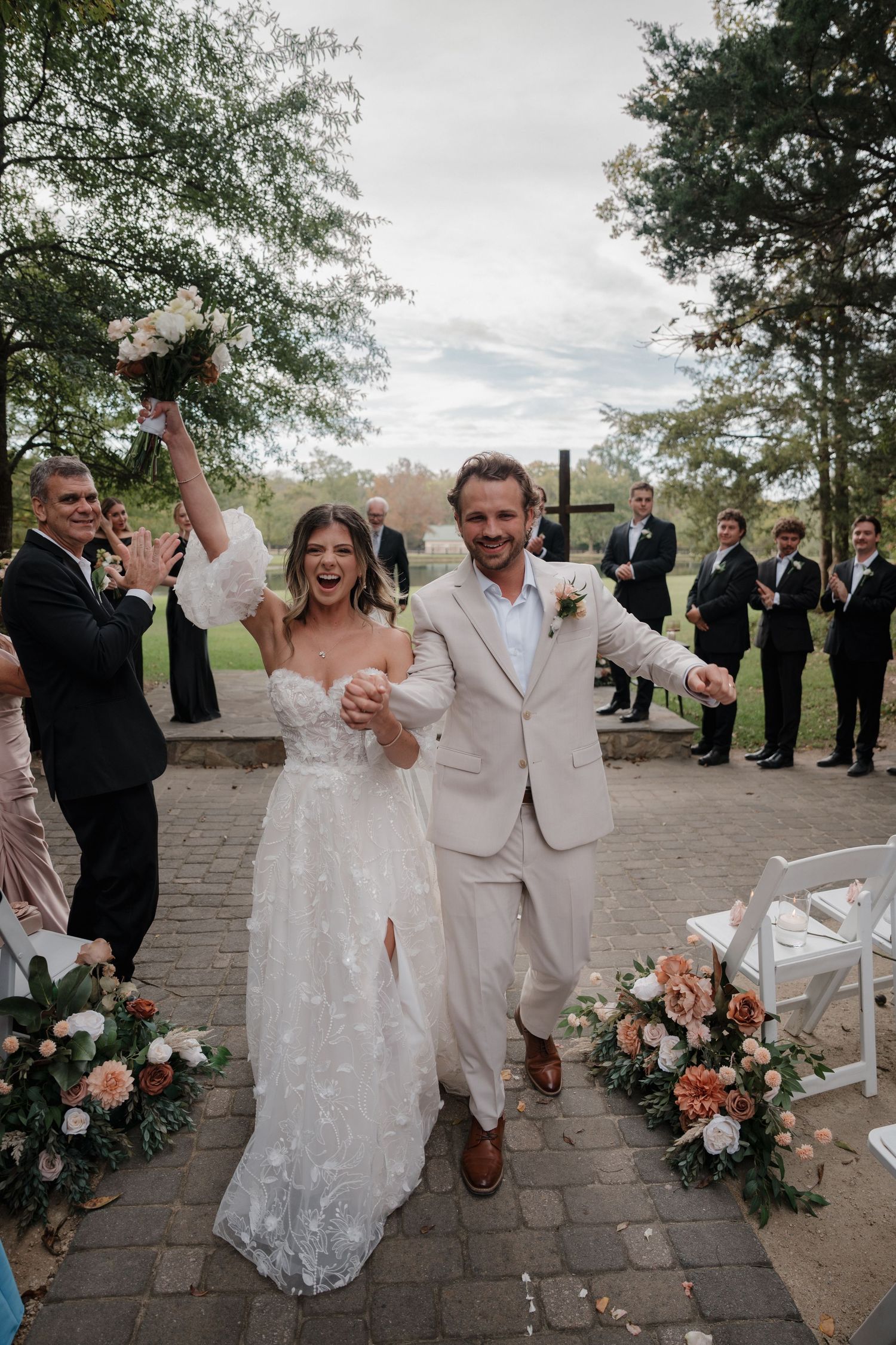 Joyful wedding couple celebrates with guests on stone patio surrounded by pink floral arrangements and greenery.
