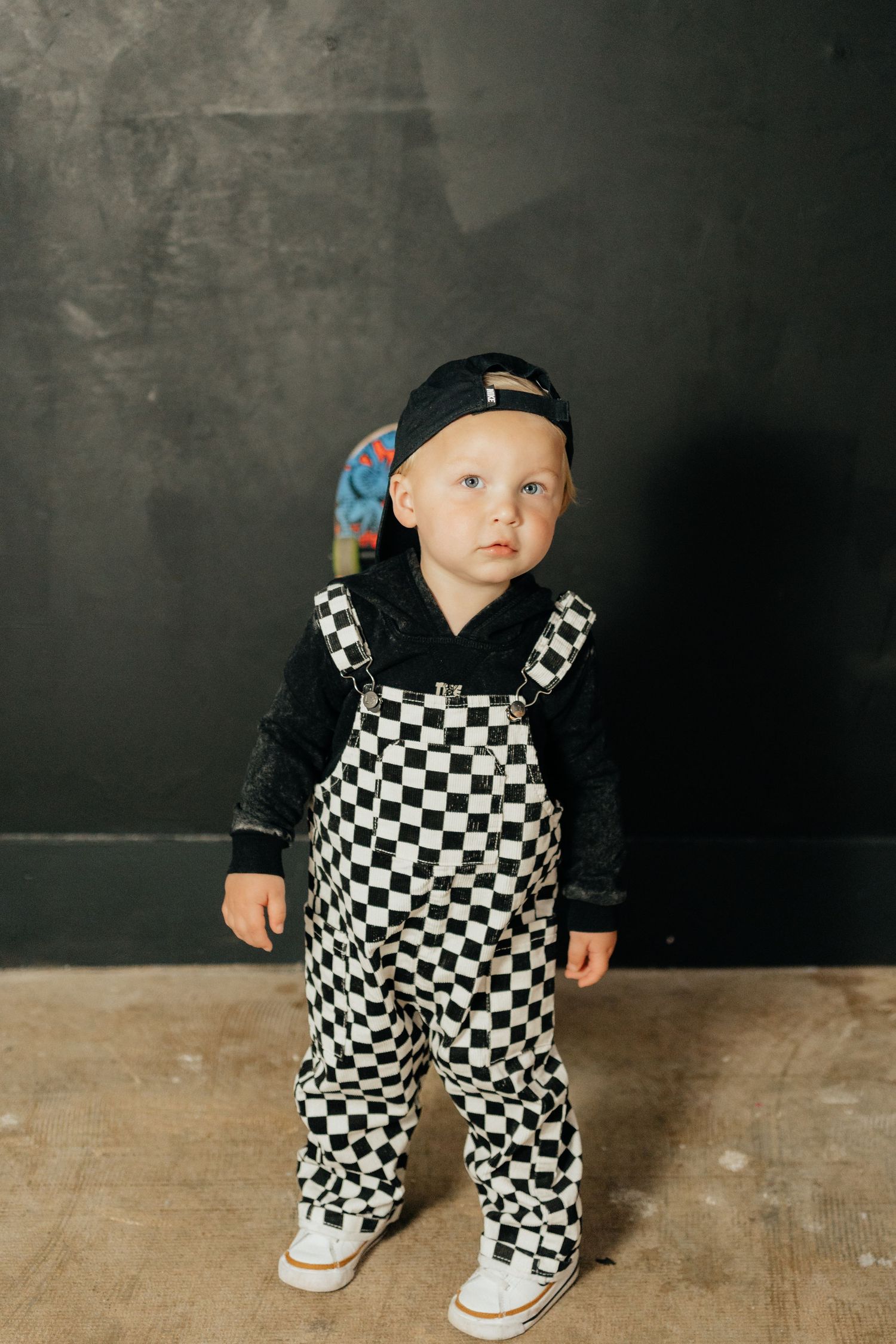 Toddler boy in black and white checkered overalls, black hoodie, and backwards cap standing against dark wall.