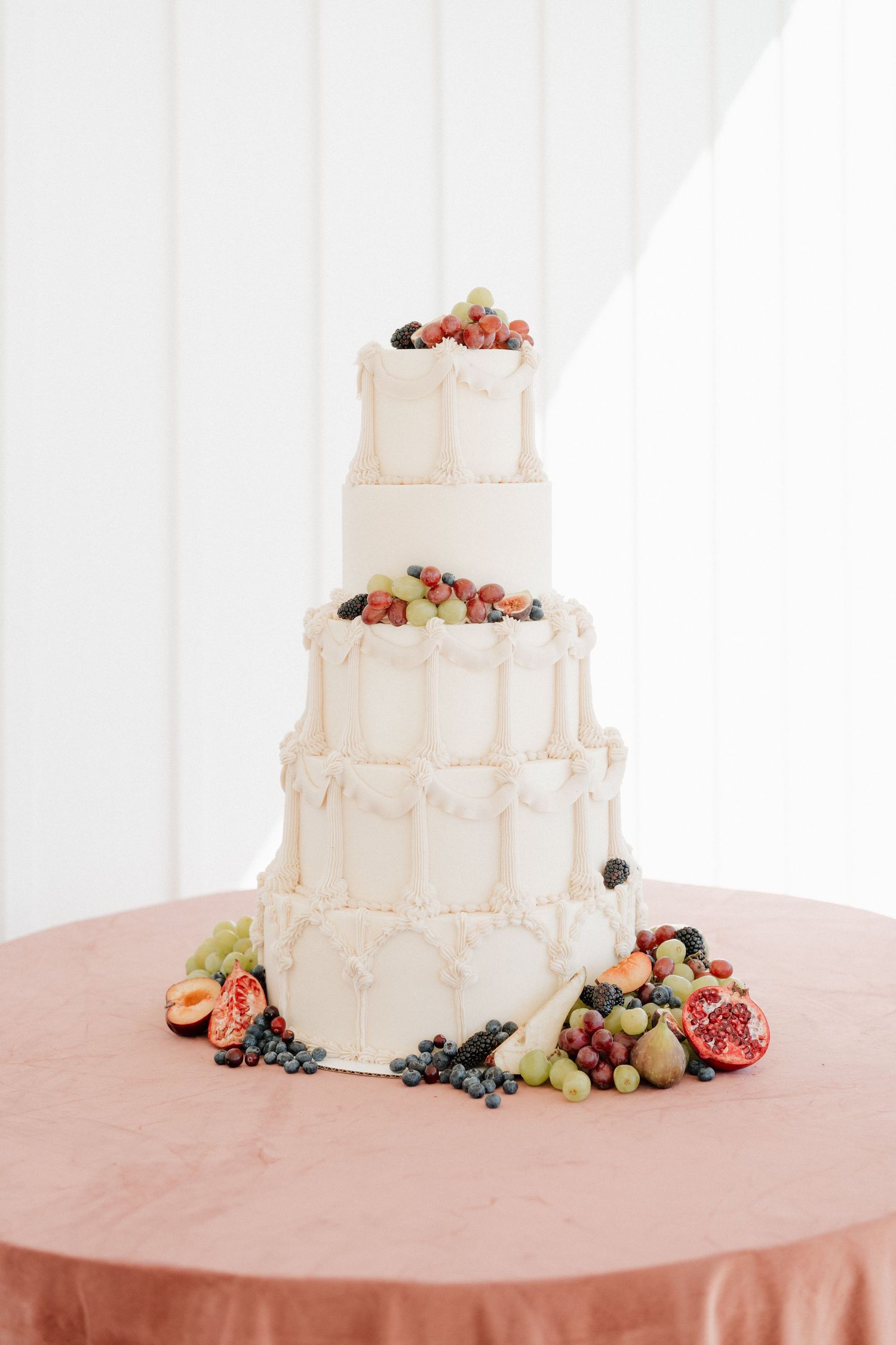 Three-tiered white wedding cake decorated with fresh berries and fruit on a pink tablecloth.