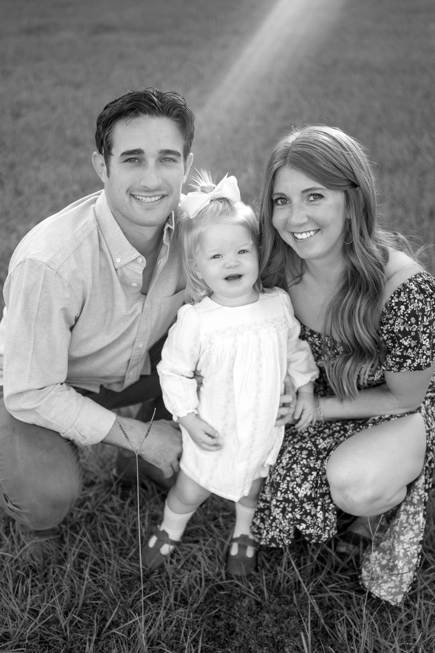 Black and white portrait of a young family with a smiling toddler wearing a white dress and bow outdoors.