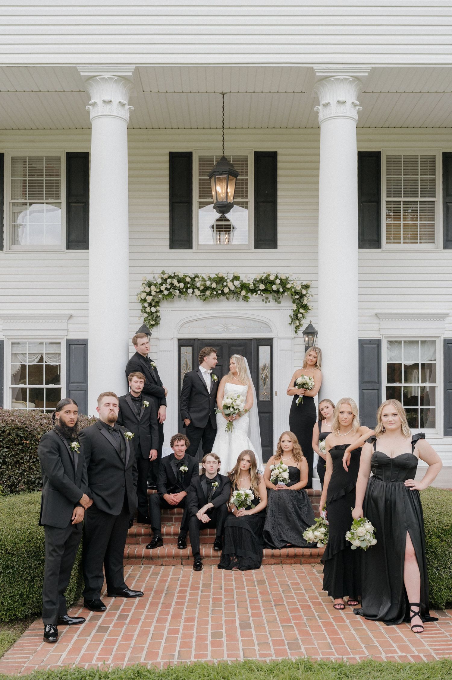 Wedding party wearing black formal attire poses on white porch steps of elegant southern home with white columns.