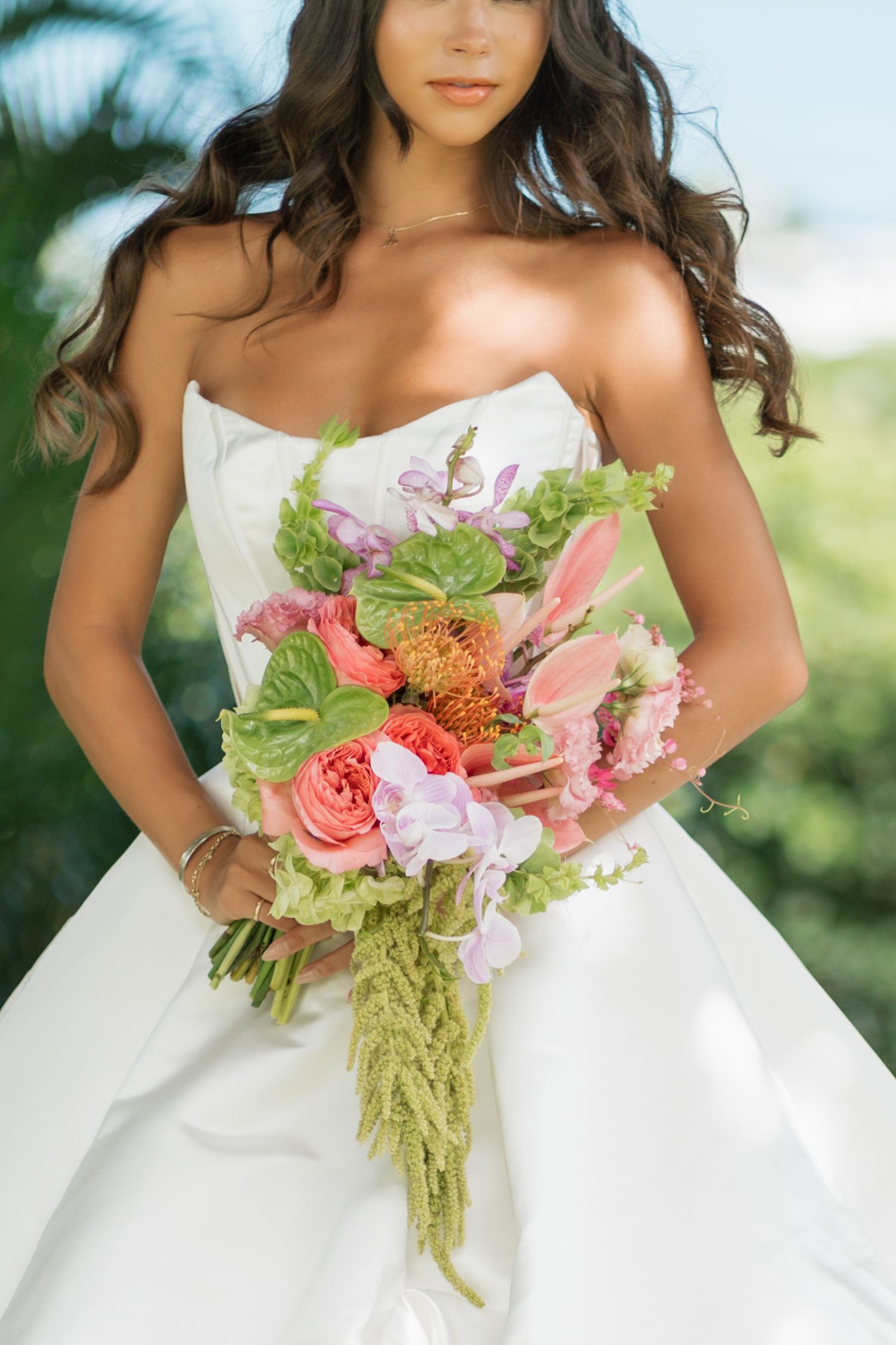 Bride in strapless white gown holding lush tropical bouquet with coral, pink, and green flowers.