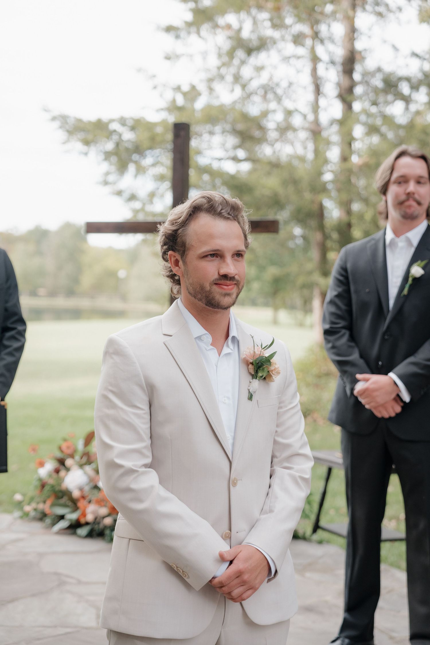 Groom in light beige suit jacket smiles during outdoor wedding ceremony with groomsmen in black suits.