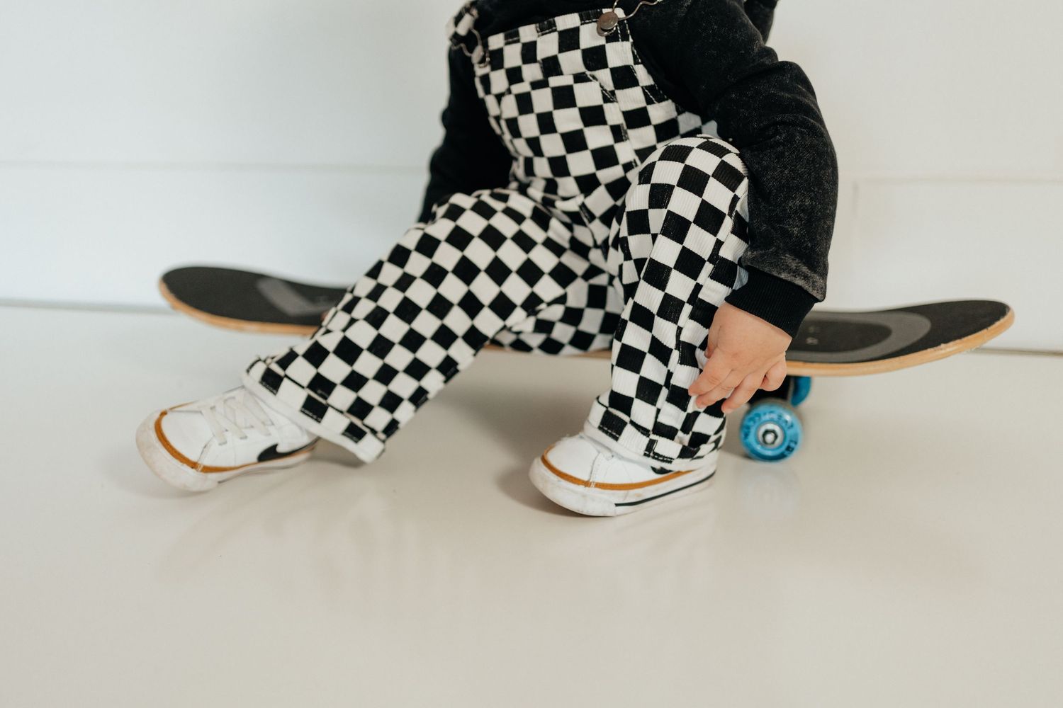Child sitting on a skateboard wearing black and white checkered pants and white sneakers.