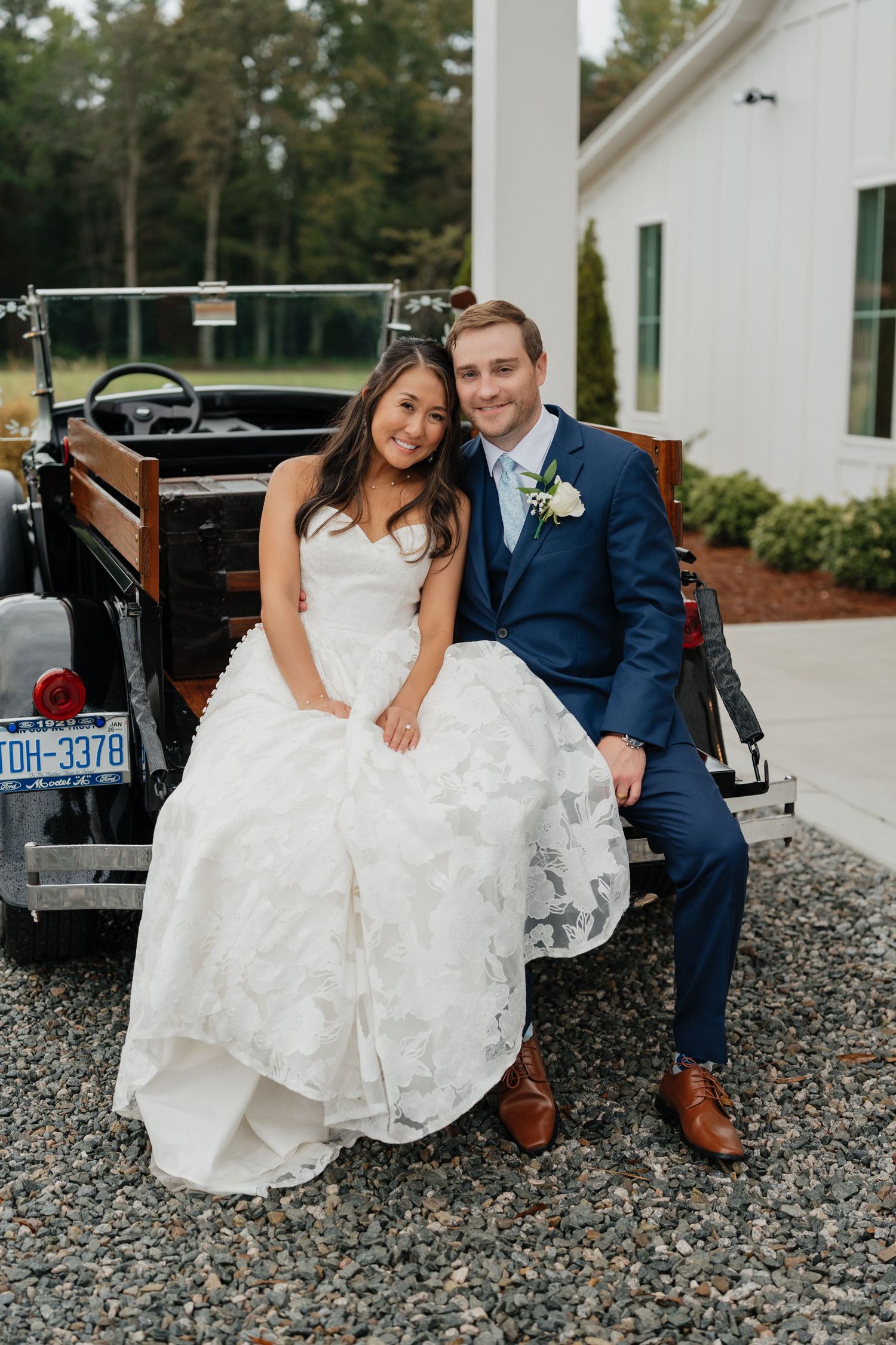 A couple in wedding attire sits together on gravel near a vintage black car in an outdoor setting.