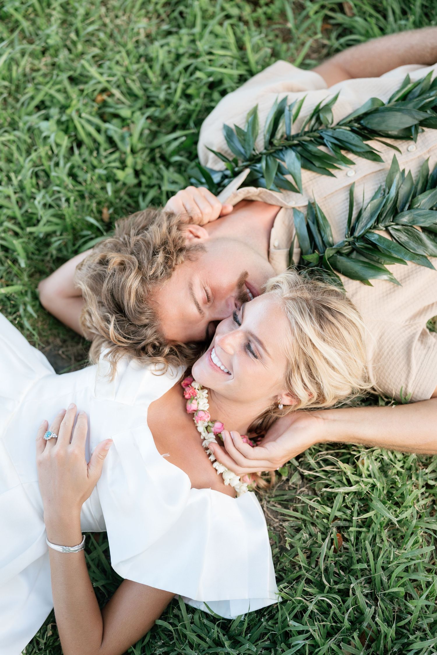 Couple in wedding attire lying on green grass, smiling, wearing floral leis and a leaf crown.