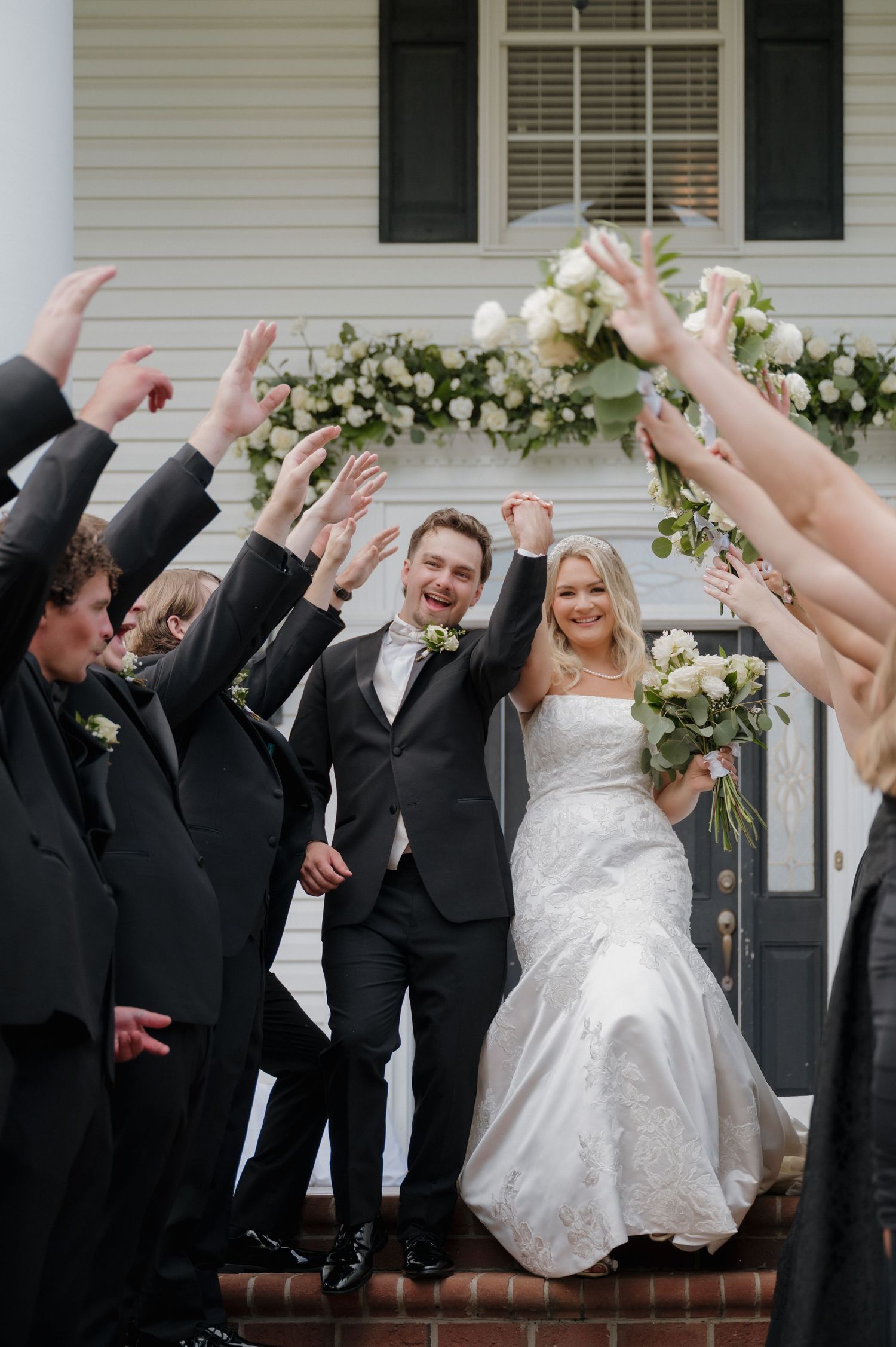 Wedding party poses for a fun jumping photo sequence in front of white house with bride and groomsmen in black suits.