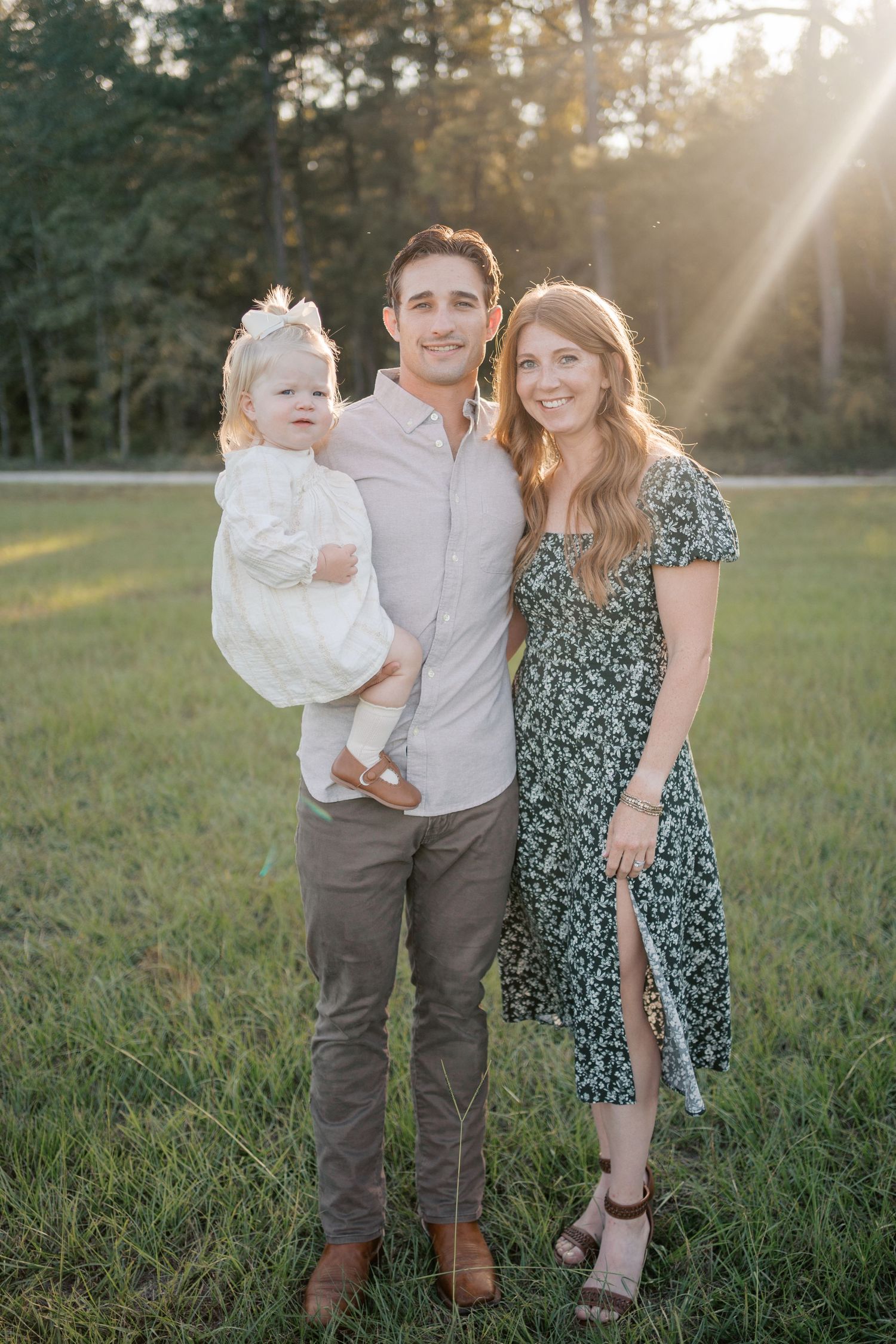 A young family poses together in a sunlit grassy field during golden hour, wearing coordinated casual outfits.