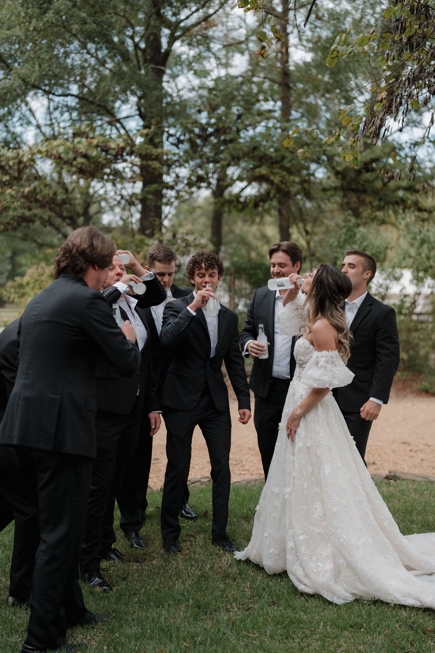 Wedding party gathered outdoors for candid group photos in a natural setting with trees in the background.