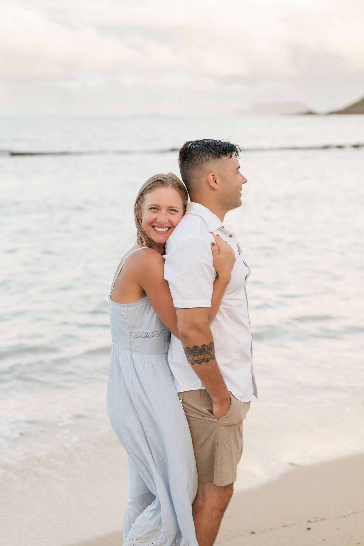 A smiling woman in a light blue dress hugs a man in a white shirt on a sandy beach with ocean waves behind them.