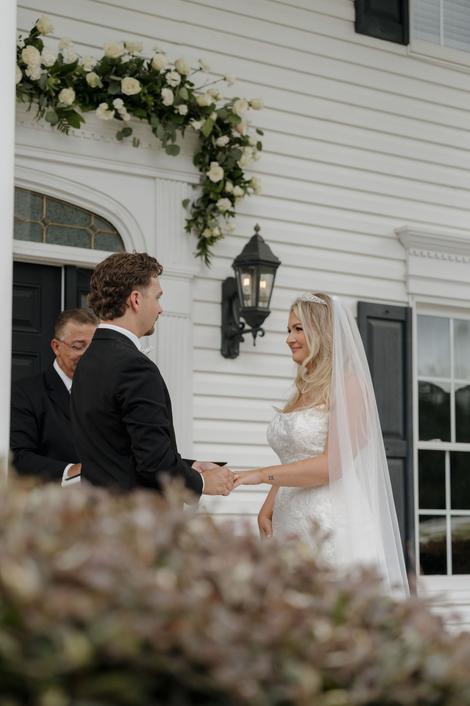 A bride and groom exchange vows in front of a white church entrance adorned with floral arrangements and a vintage lantern.