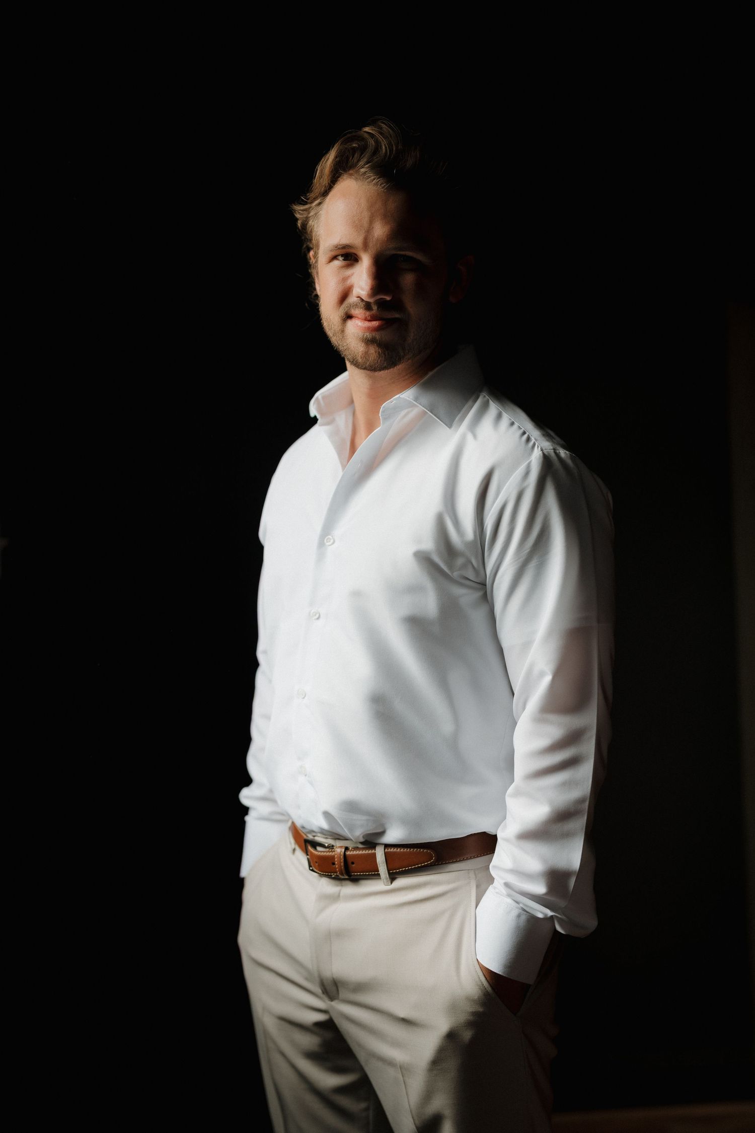 A person in a crisp white dress shirt and khaki pants poses against a dark background with dramatic lighting.