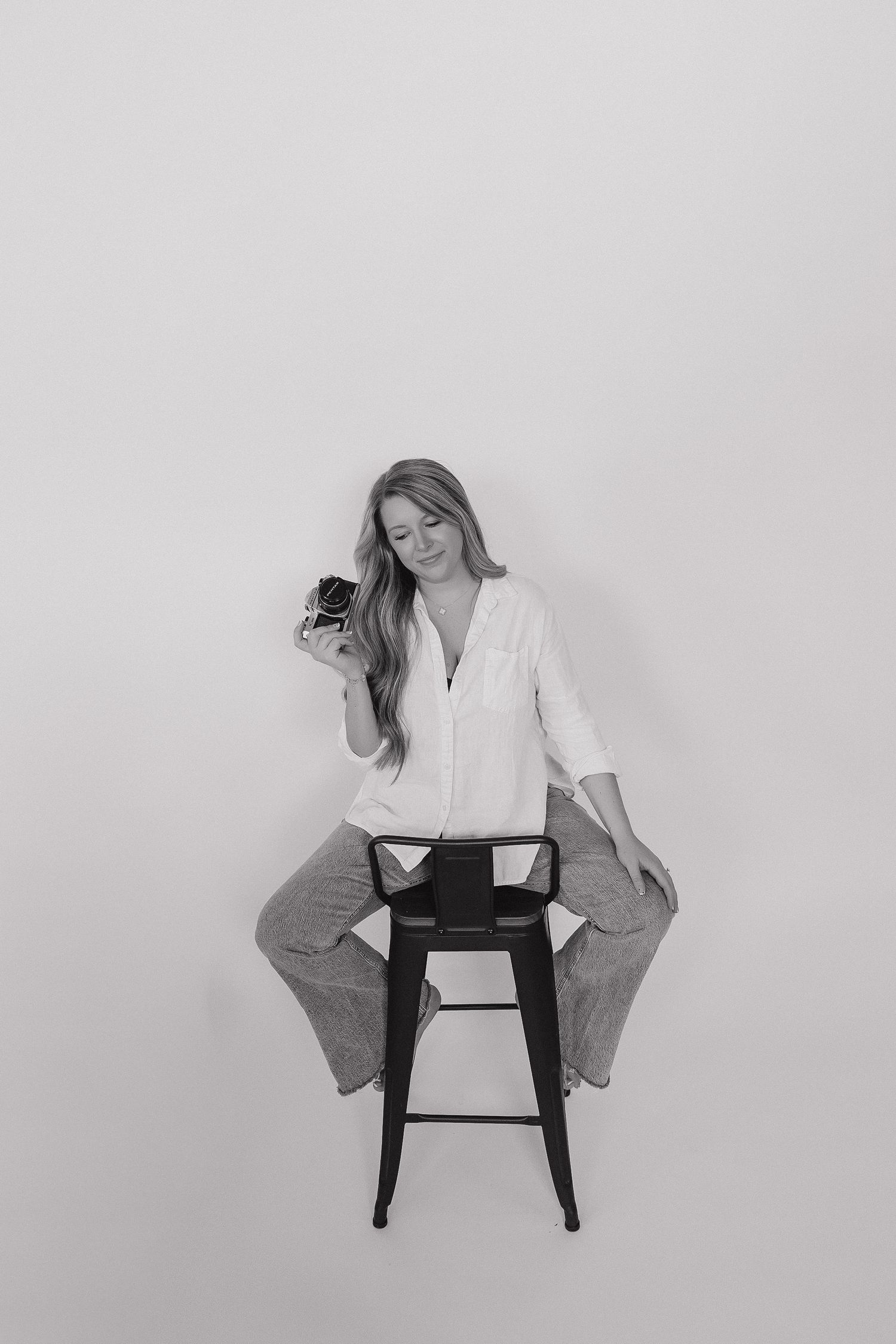 Black and white photo of a woman sitting on a stool holding a vintage camera, wearing a white shirt and jeans.