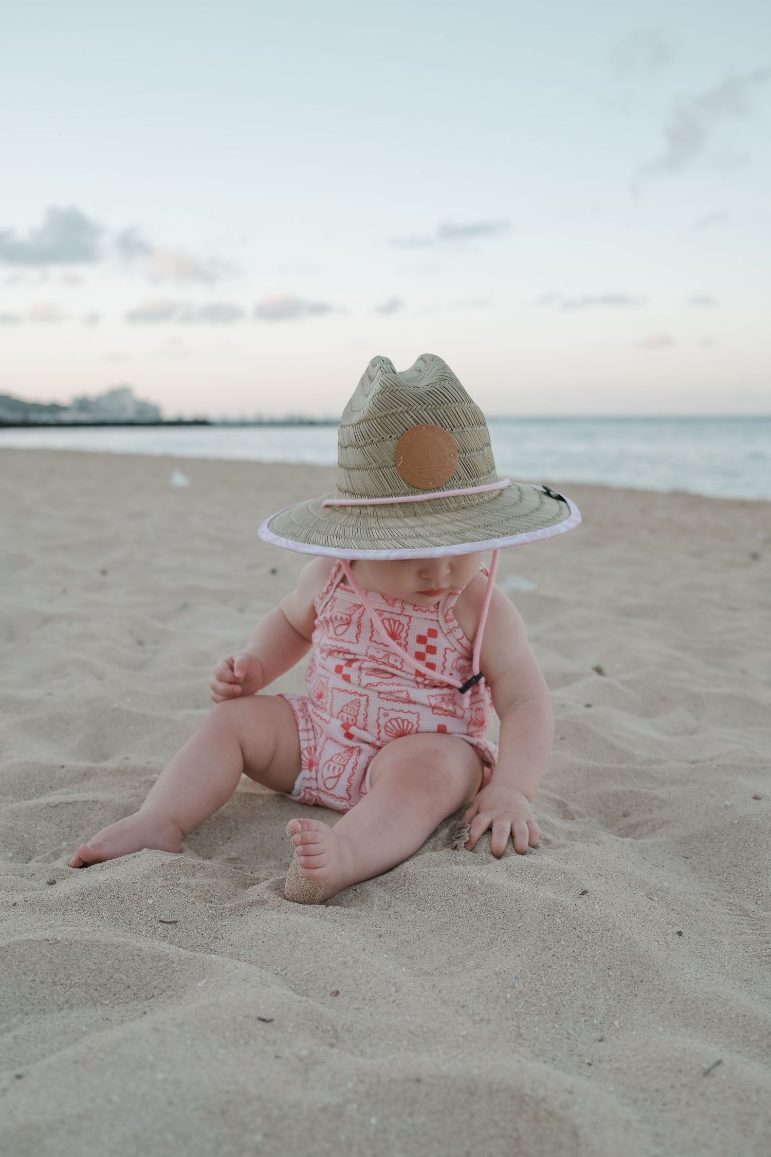 Adorable baby wearing a straw sun hat sitting on sandy beach at sunset near the ocean.