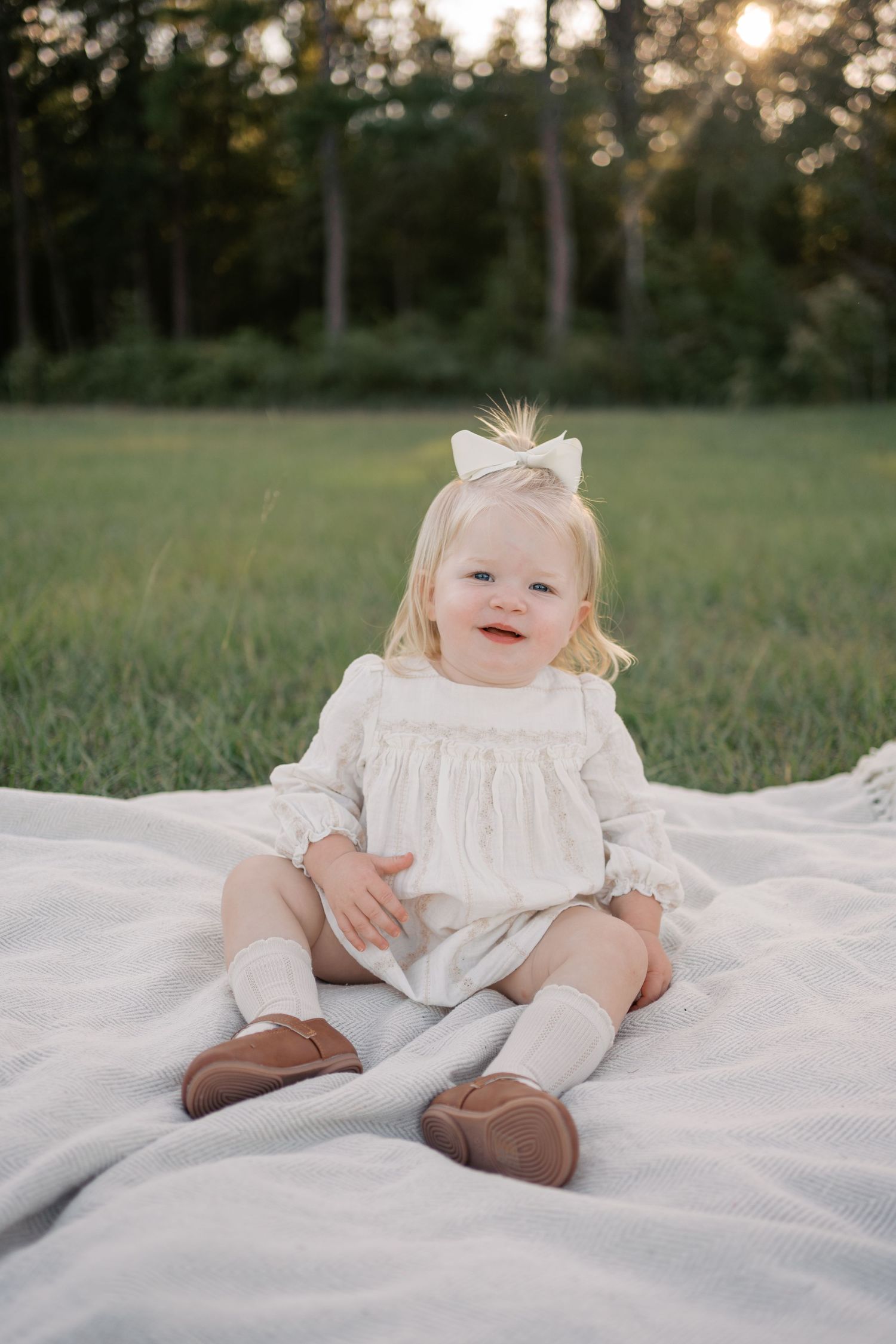 A baby in a white dress sits on a white blanket outdoors during golden hour with trees in the background.