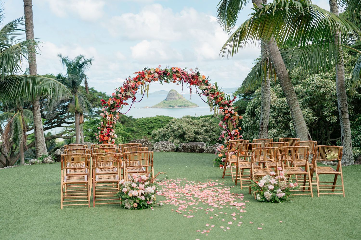 Outdoor wedding ceremony setup with floral arch, bamboo chairs, and rose petal aisle on tropical lawn with palm trees.