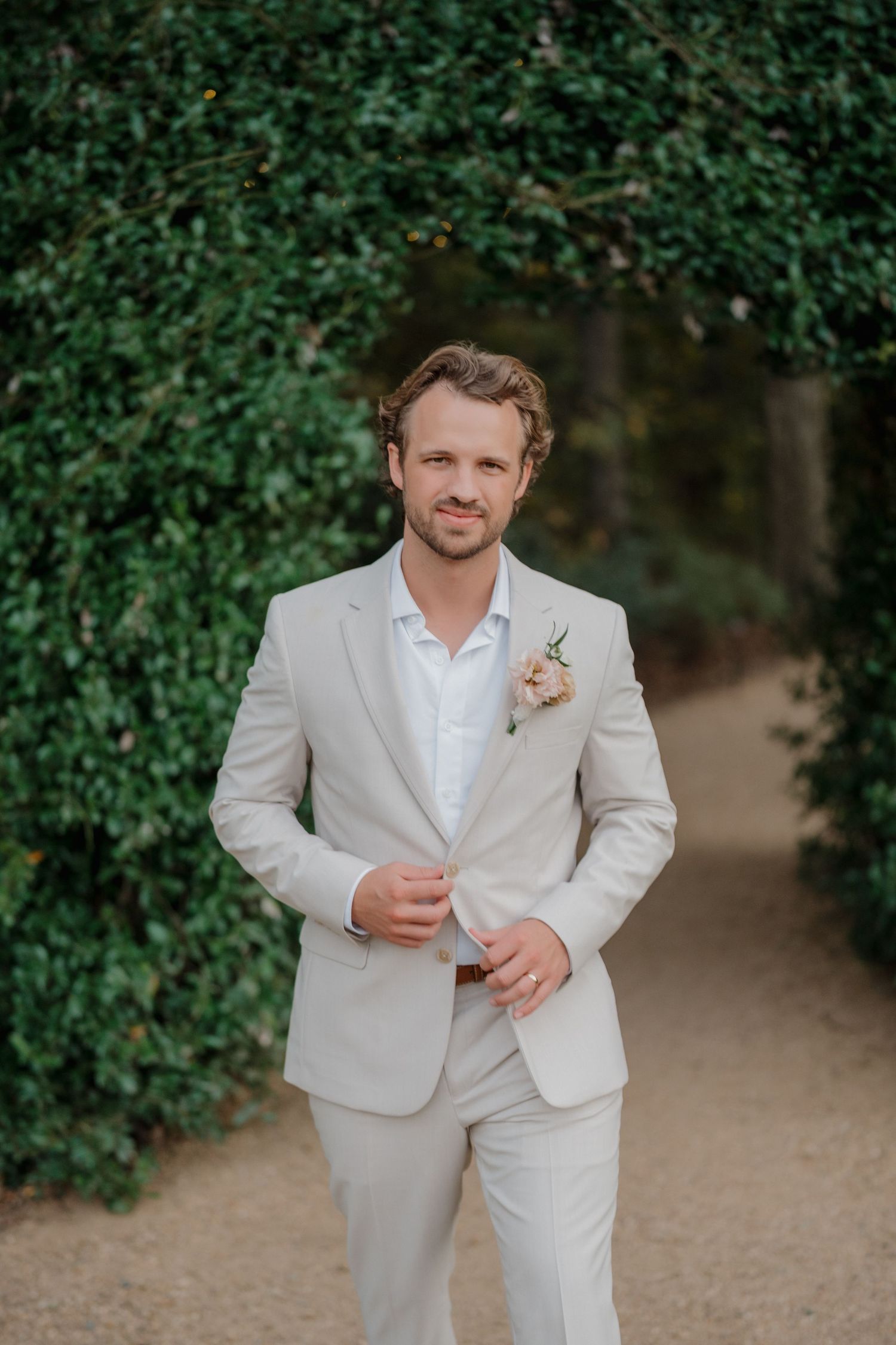 A groom in a light beige suit and white shirt stands in front of a lush green hedge on a garden path.