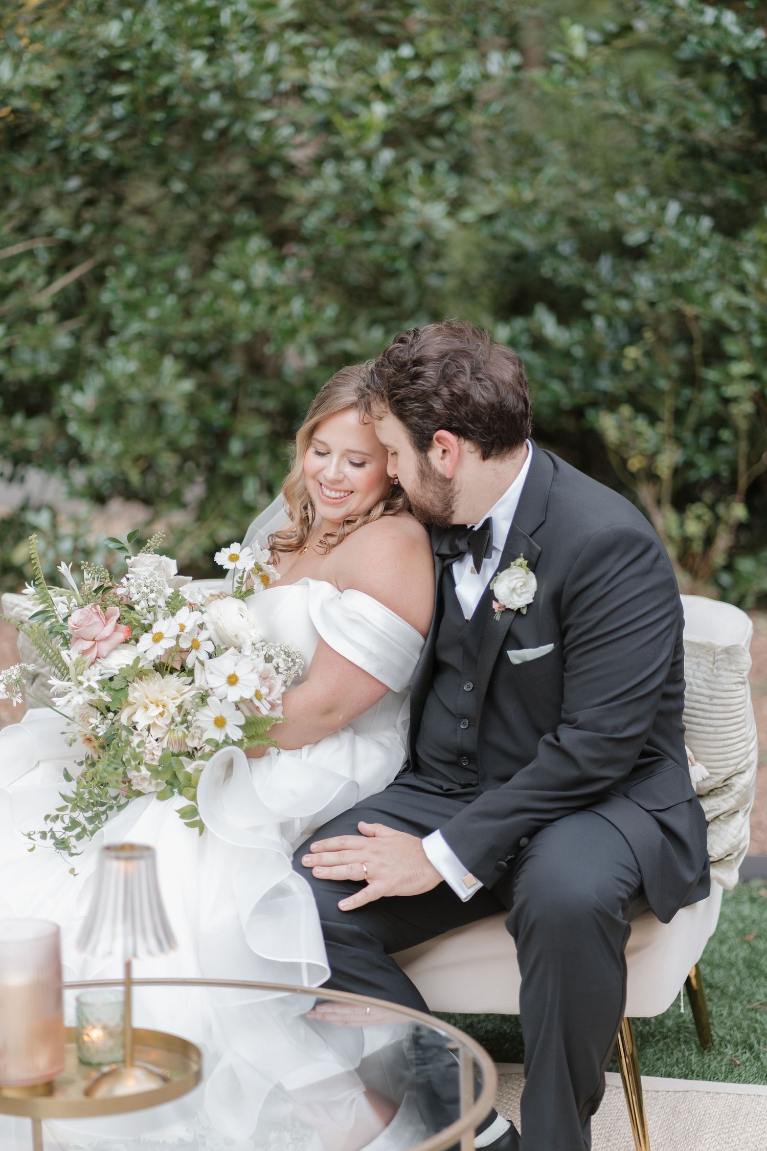 A romantic wedding couple shares tender moments while seated outdoors surrounded by greenery and floral arrangements.