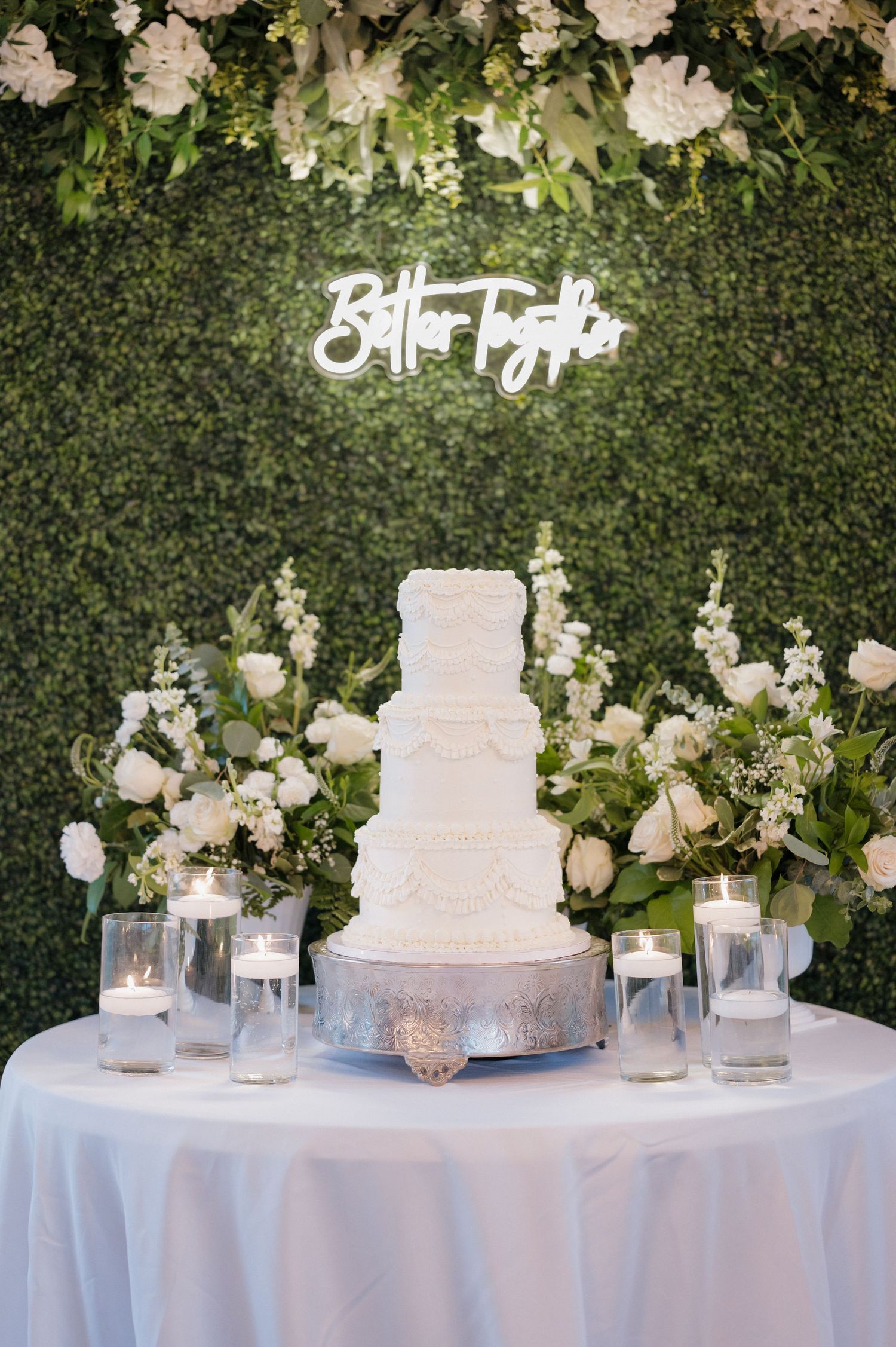 Elegant white wedding cake displayed on round table with floral decorations and greenery wall backdrop featuring illuminated sign.
