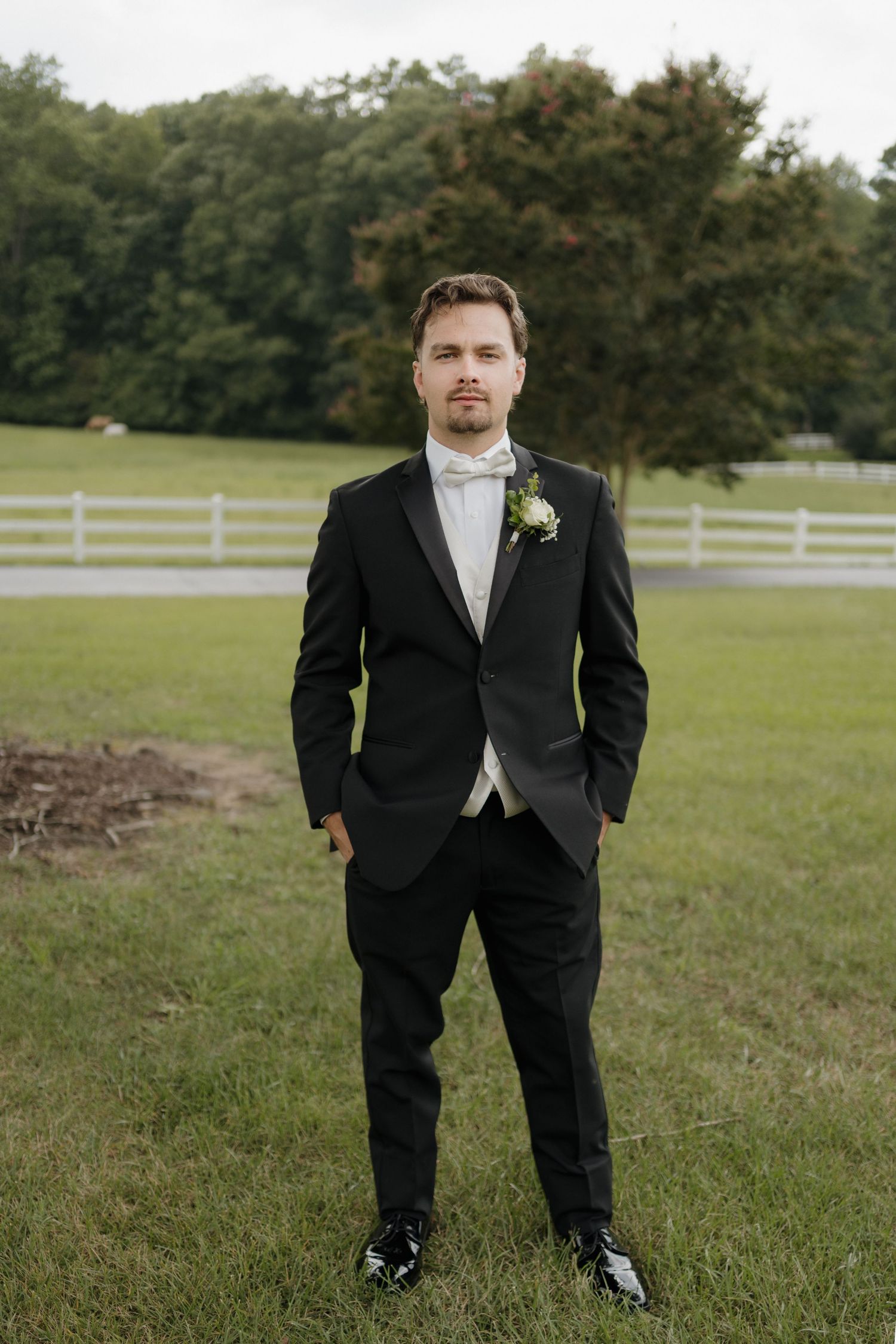 A groom in a black tuxedo stands in a grassy field with white fencing and trees in the background.